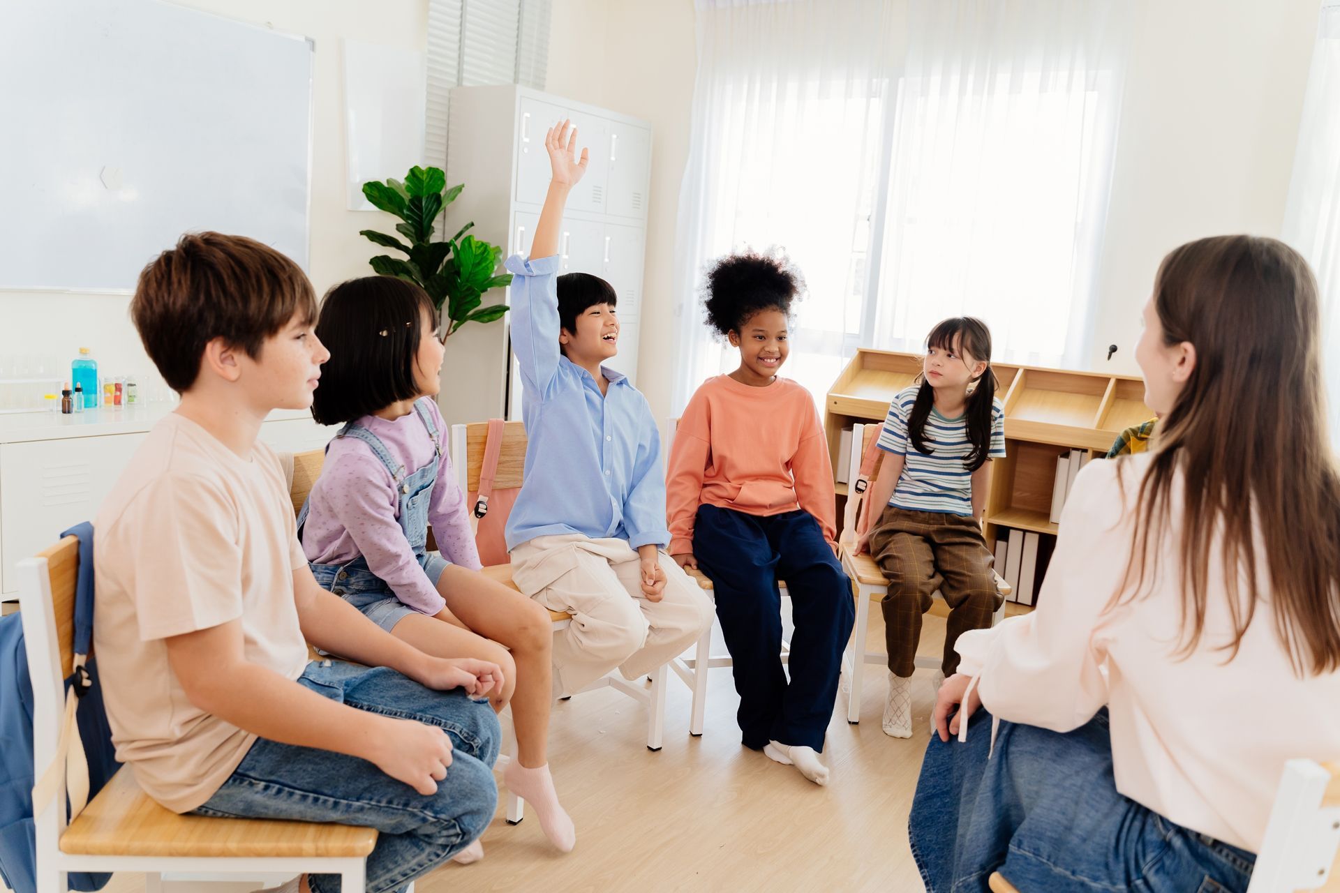 A diverse group of children sit in a circle in a classroom, with one child raising their hand to speak to an instructor.
