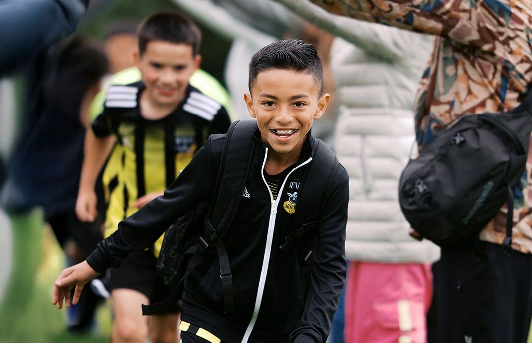 Boy in black jacket smiling, running towards camera on a field; another boy in a soccer uniform in background.