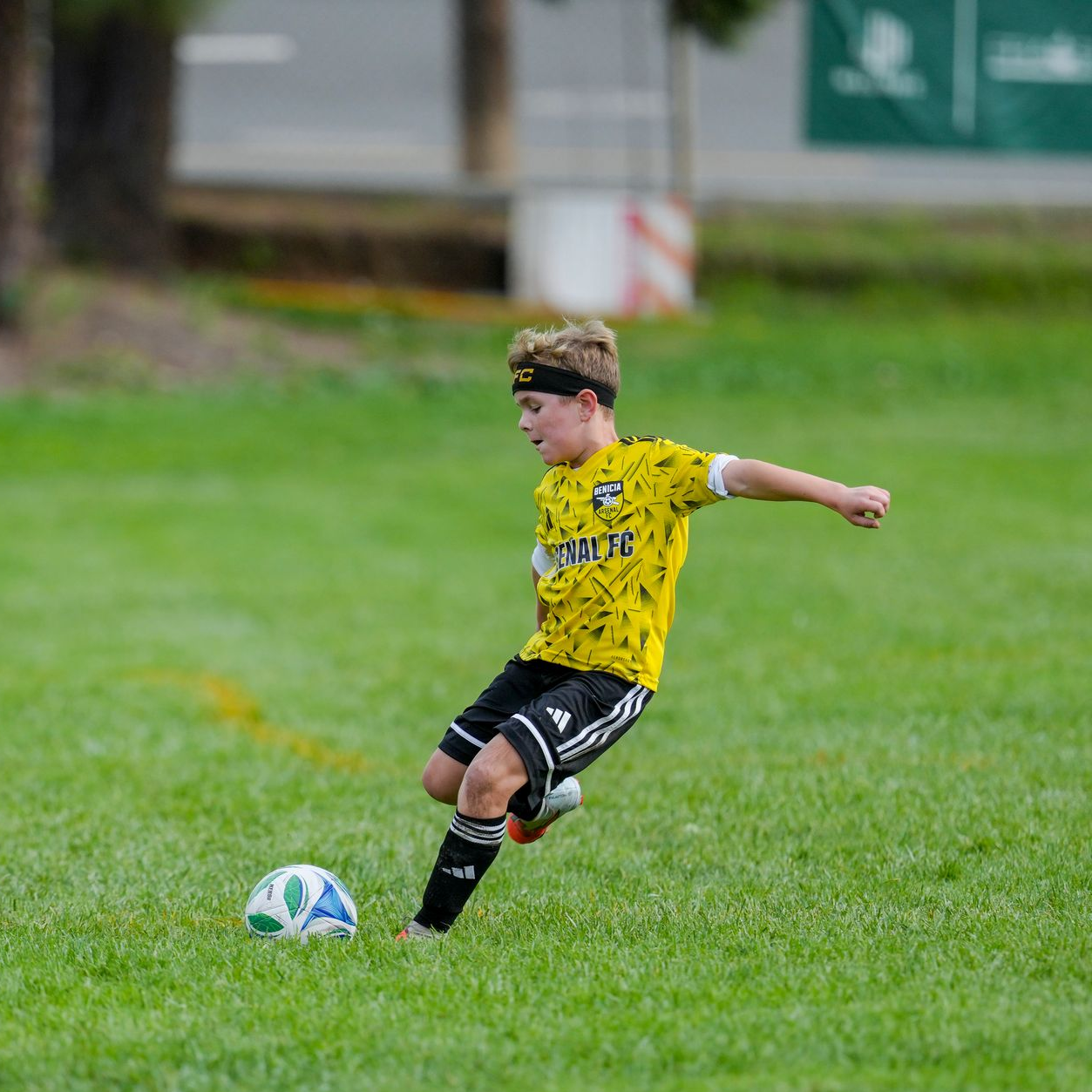 Boy in white soccer jersey kicks a soccer ball on a green field.