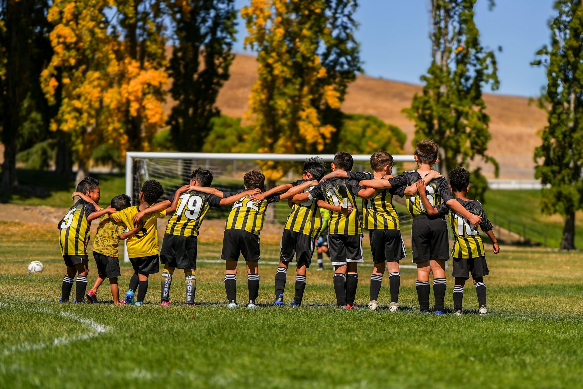 Soccer team huddles on a green field. Players in black and yellow uniforms stand with arms around each other.