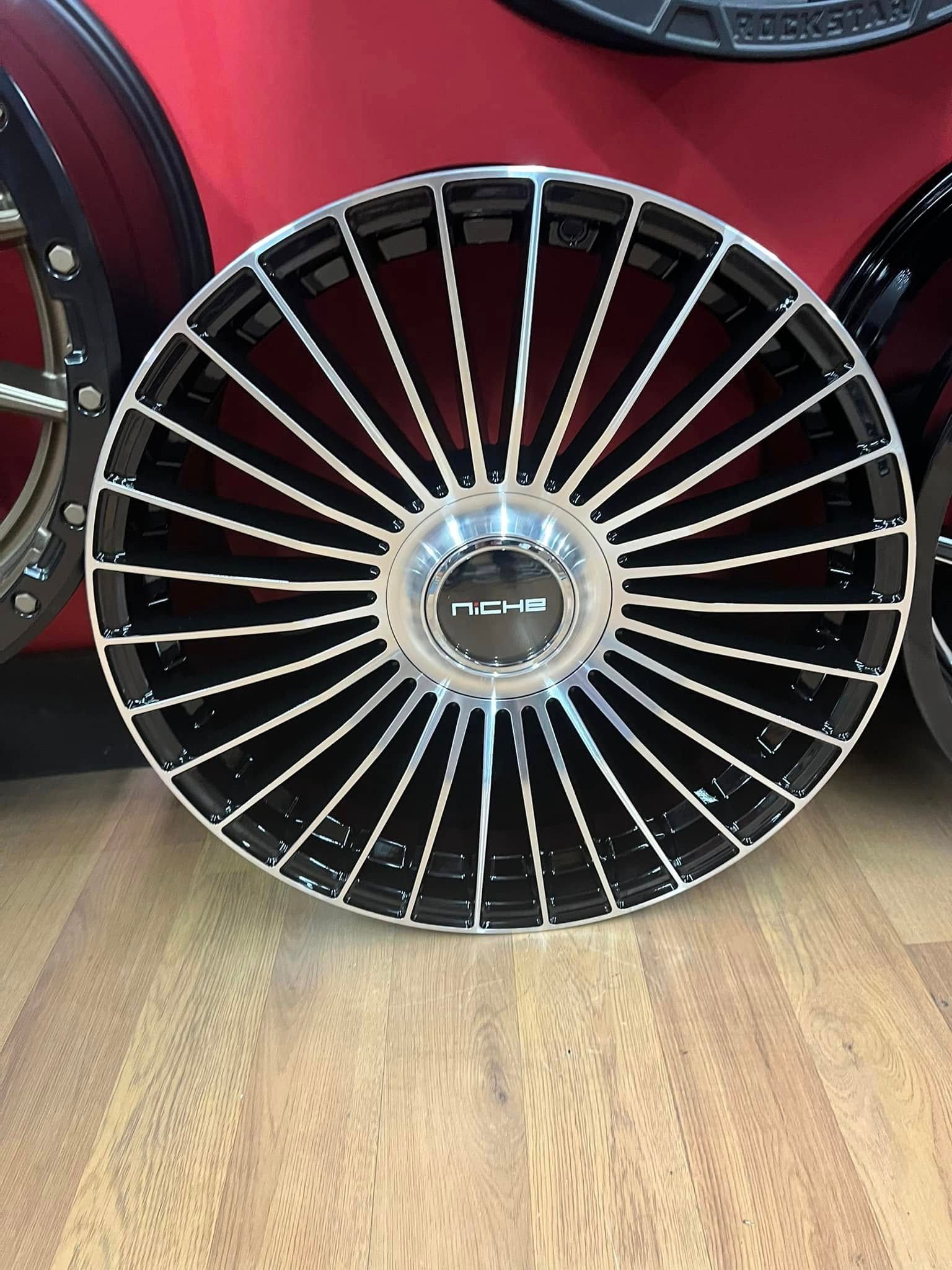 Black and silver car rim with radial spokes, set against a red background and wooden floor.