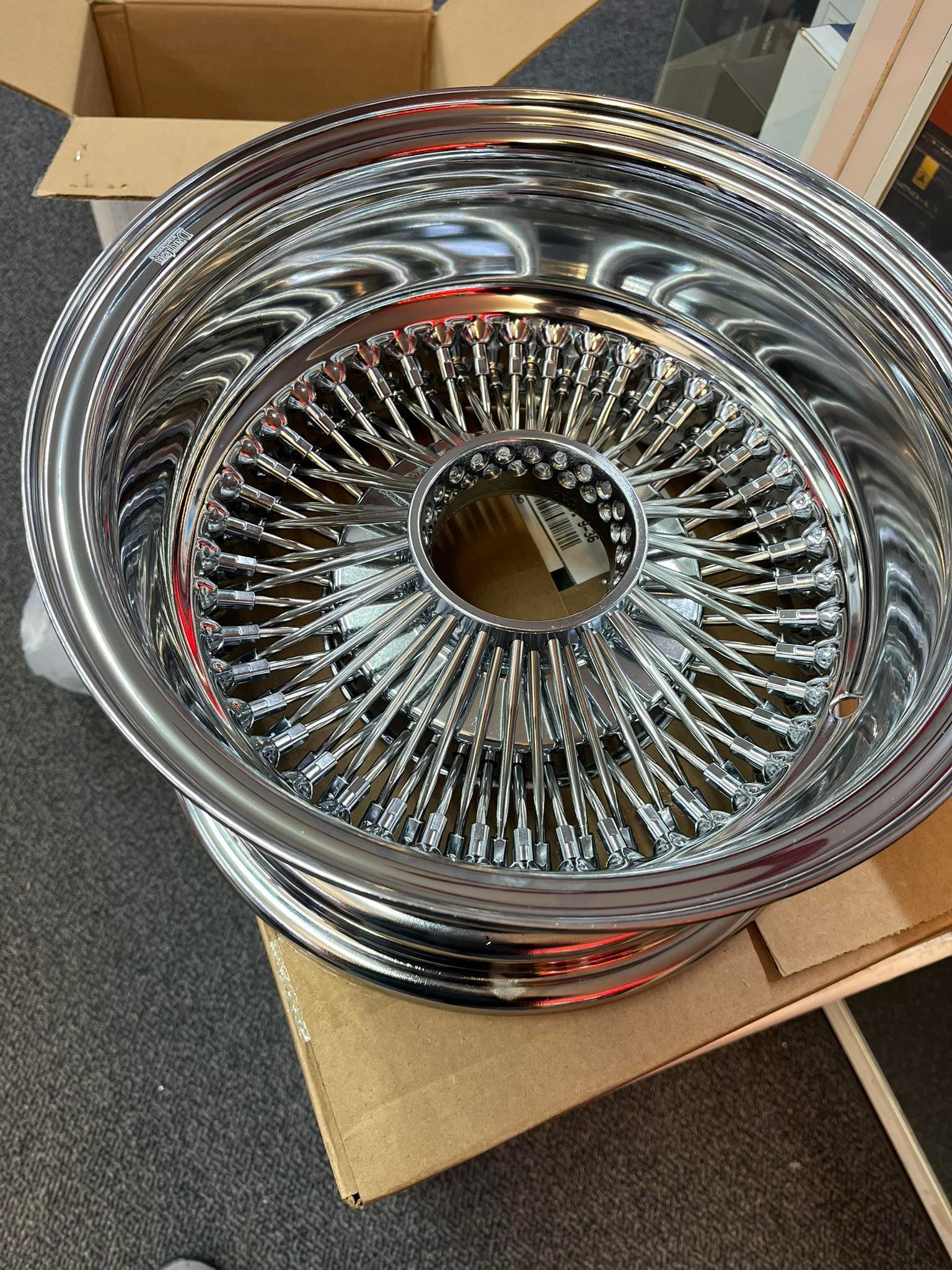 Shiny chrome wire wheel, circular design, with central hub, on a cardboard box.