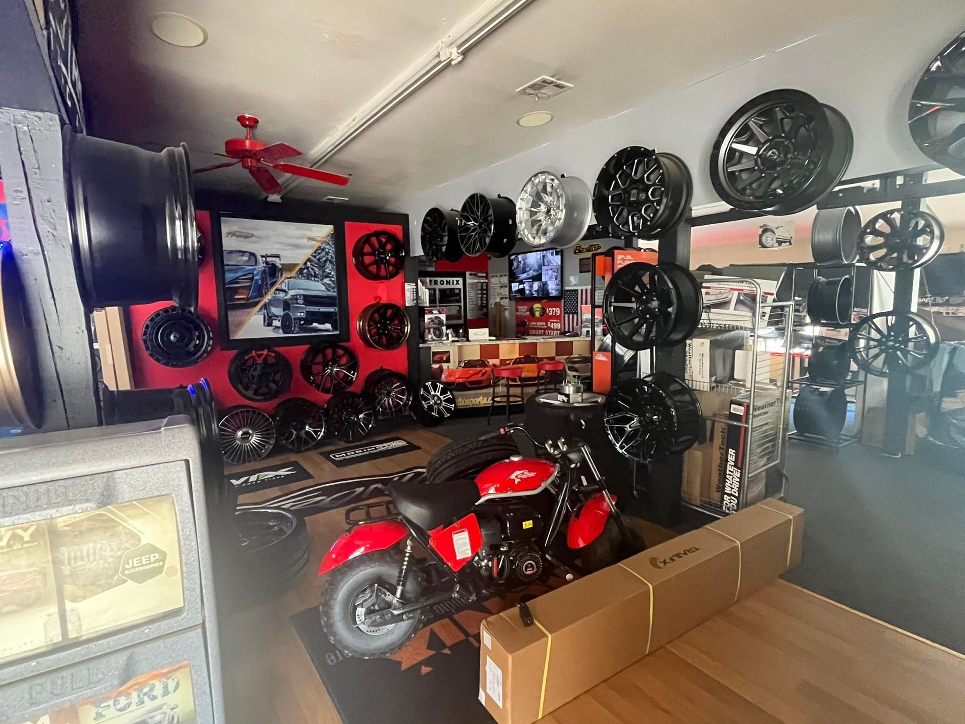 Interior of a tire shop showcasing various wheels. A red mini motorcycle sits center with boxes nearby.