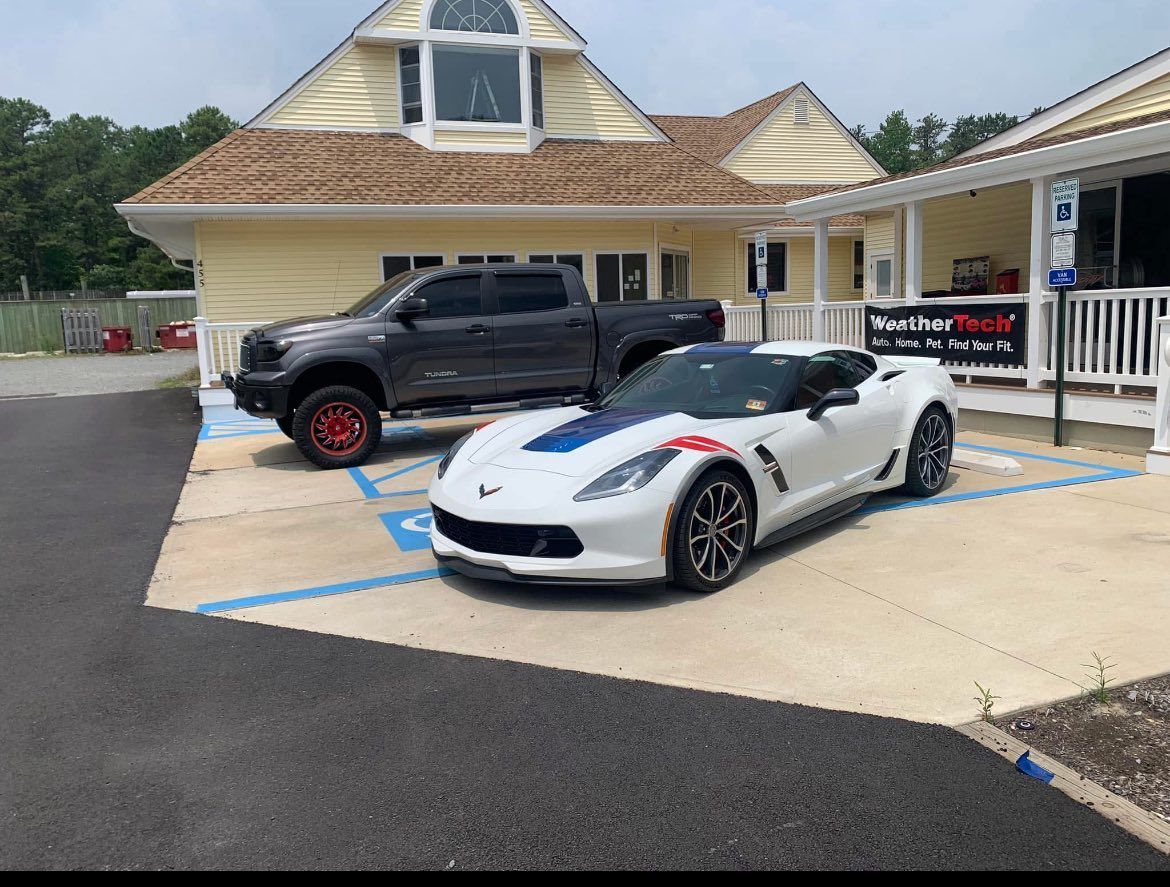 White sports car with blue stripe and lifted gray truck parked outside a yellow building.
