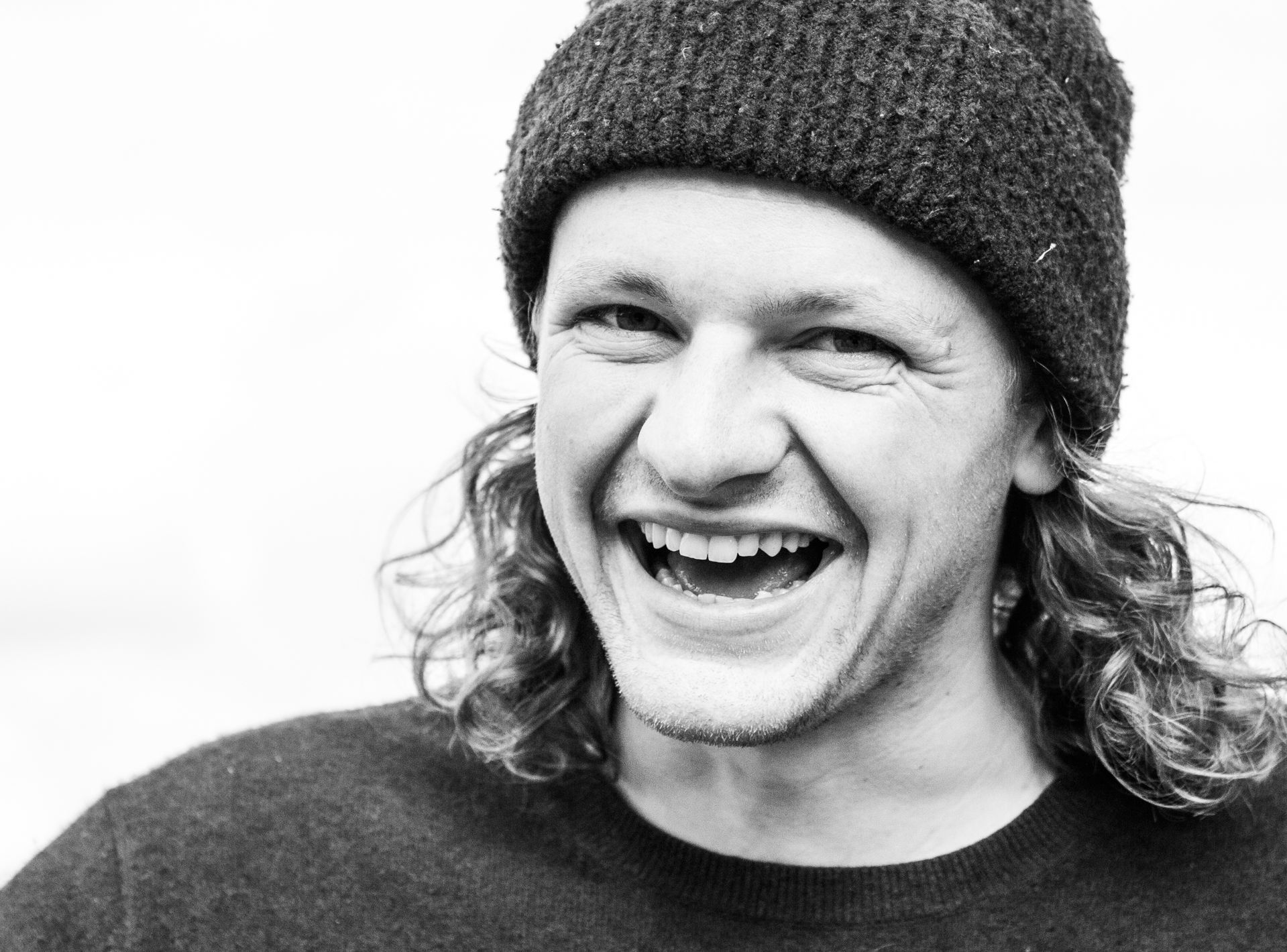 Schwarzweißfoto eines jungen englischen Mannes Surfers mit langen Locken und Mütze der in die Kamera lächelt