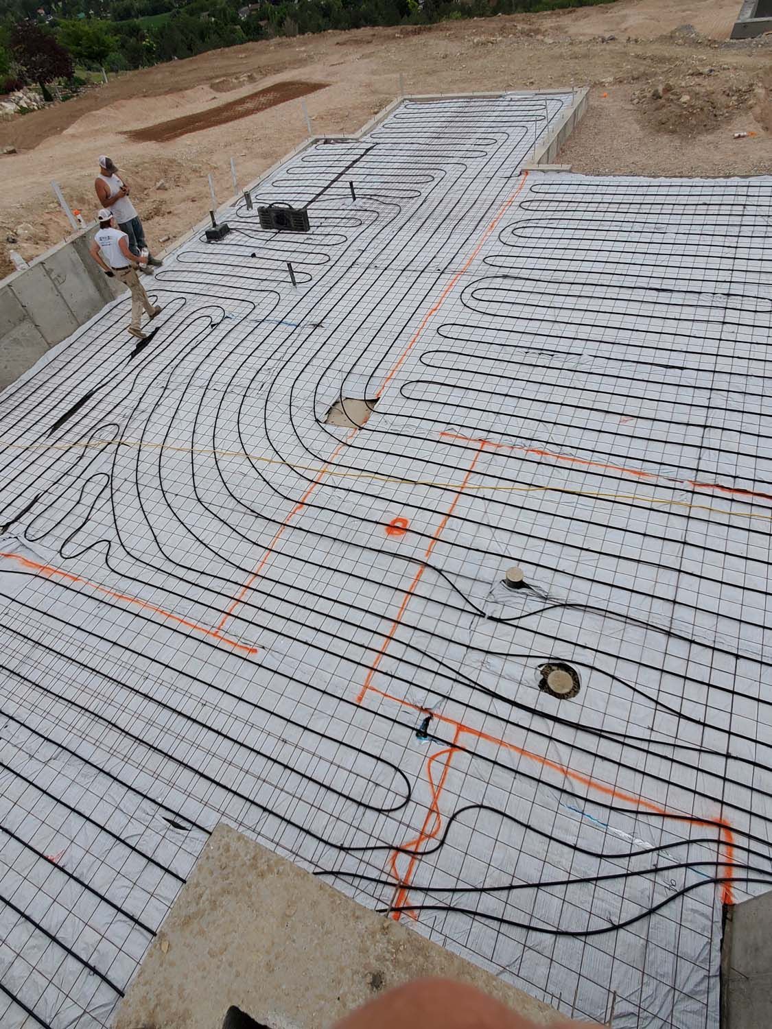 Workers installing black tubing for radiant floor heating on a white foam insulation base, with orange layout markings.