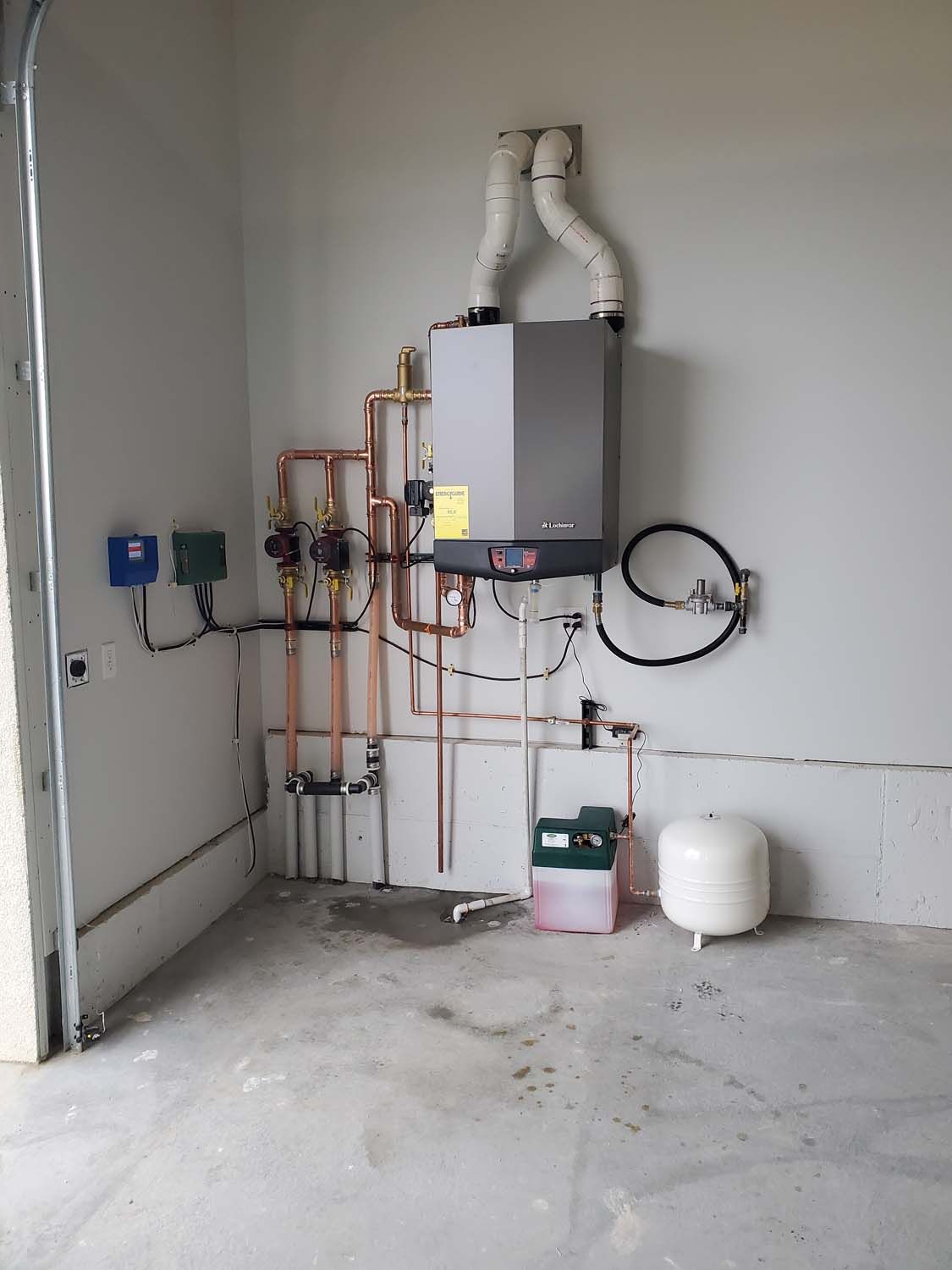 Water heater and related plumbing on a garage wall, including pipes, vents, and an expansion tank.