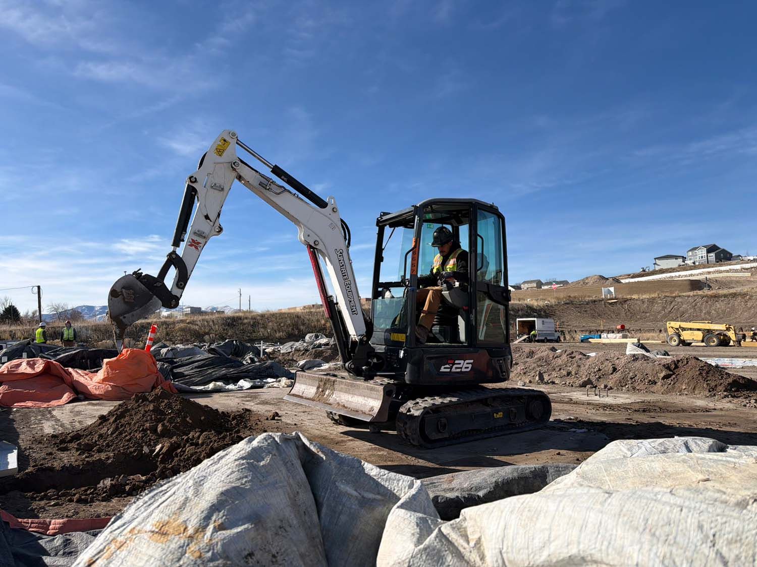 Mini excavator digging on a construction site; clear sky, worker in cab, dirt piles.