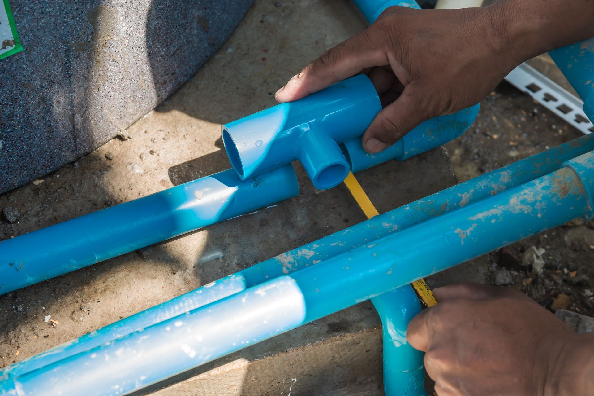 Hands assembling blue PVC pipes with a tee fitting on a concrete surface.