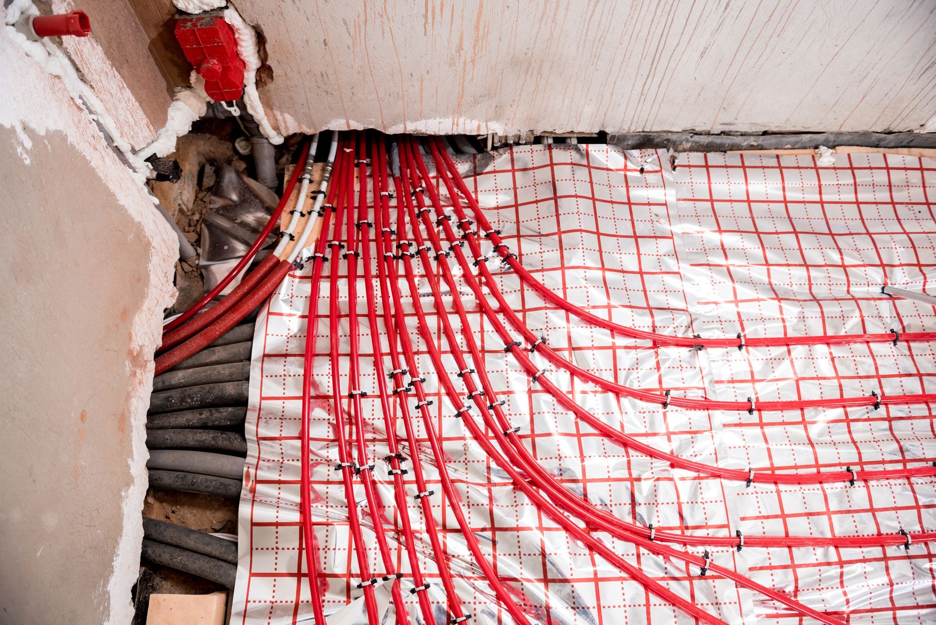 Red radiant heating tubes emerging from a wall, laid over a foil-backed grid, inside a building.