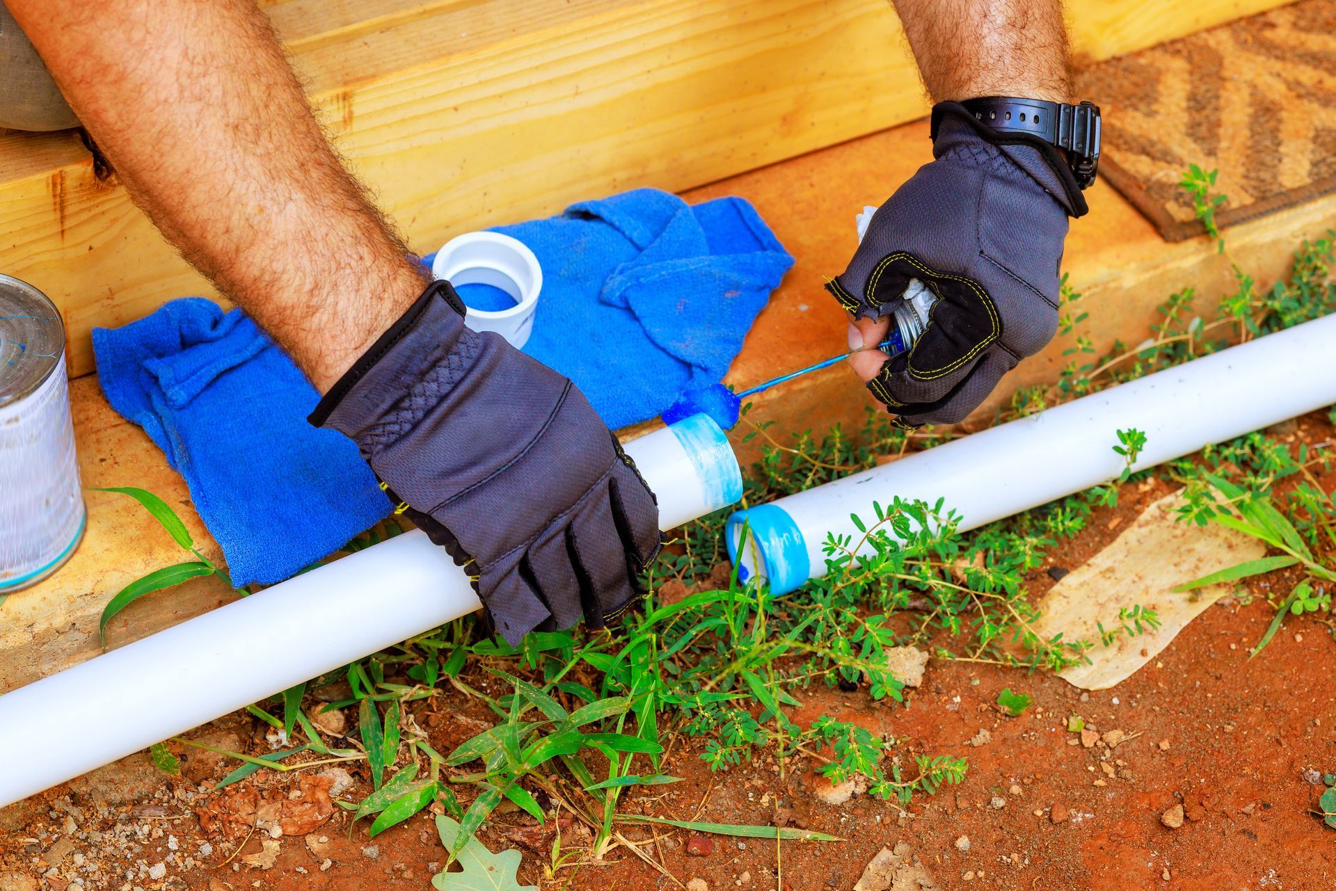 Person connecting PVC pipes outdoors, wearing gloves; blue cloth, solvent can nearby.