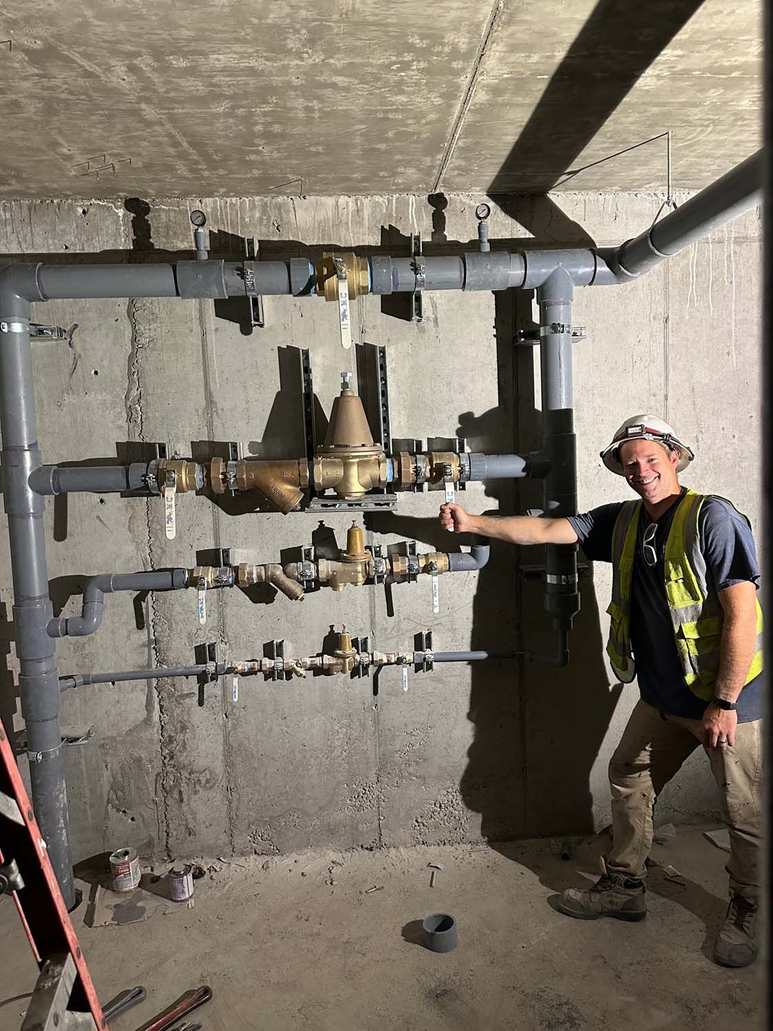 Plumber pointing to water pipes on a concrete wall. He wears a hard hat and safety vest.