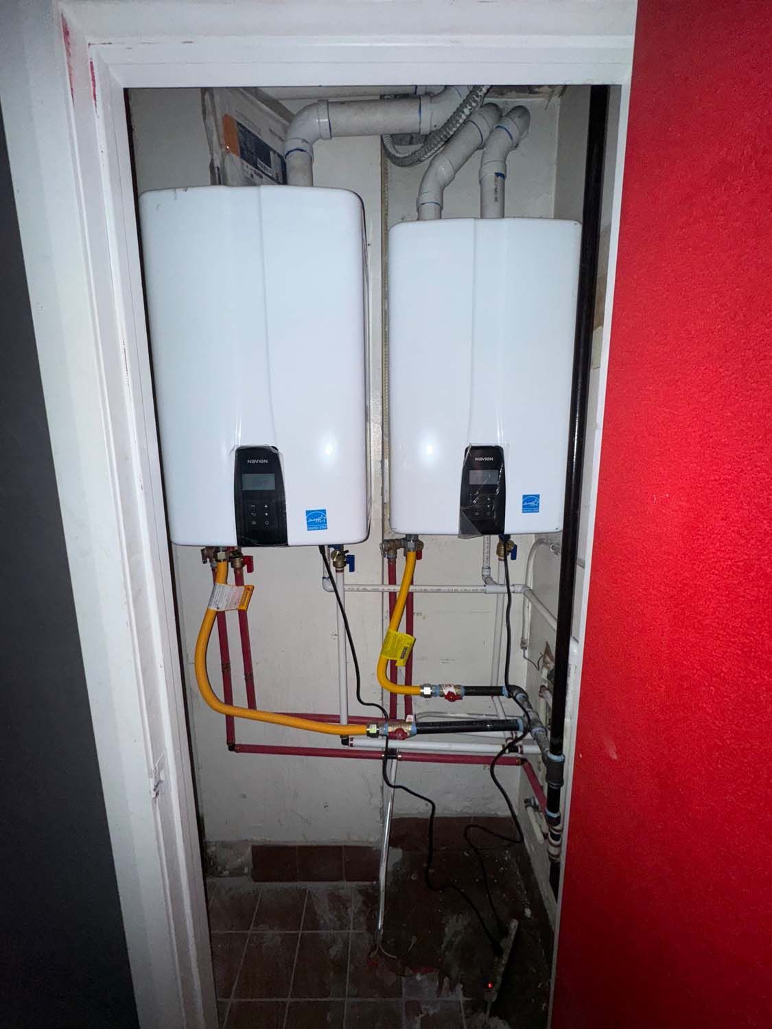Two white water heaters installed in a closet with yellow gas lines and exposed pipes.