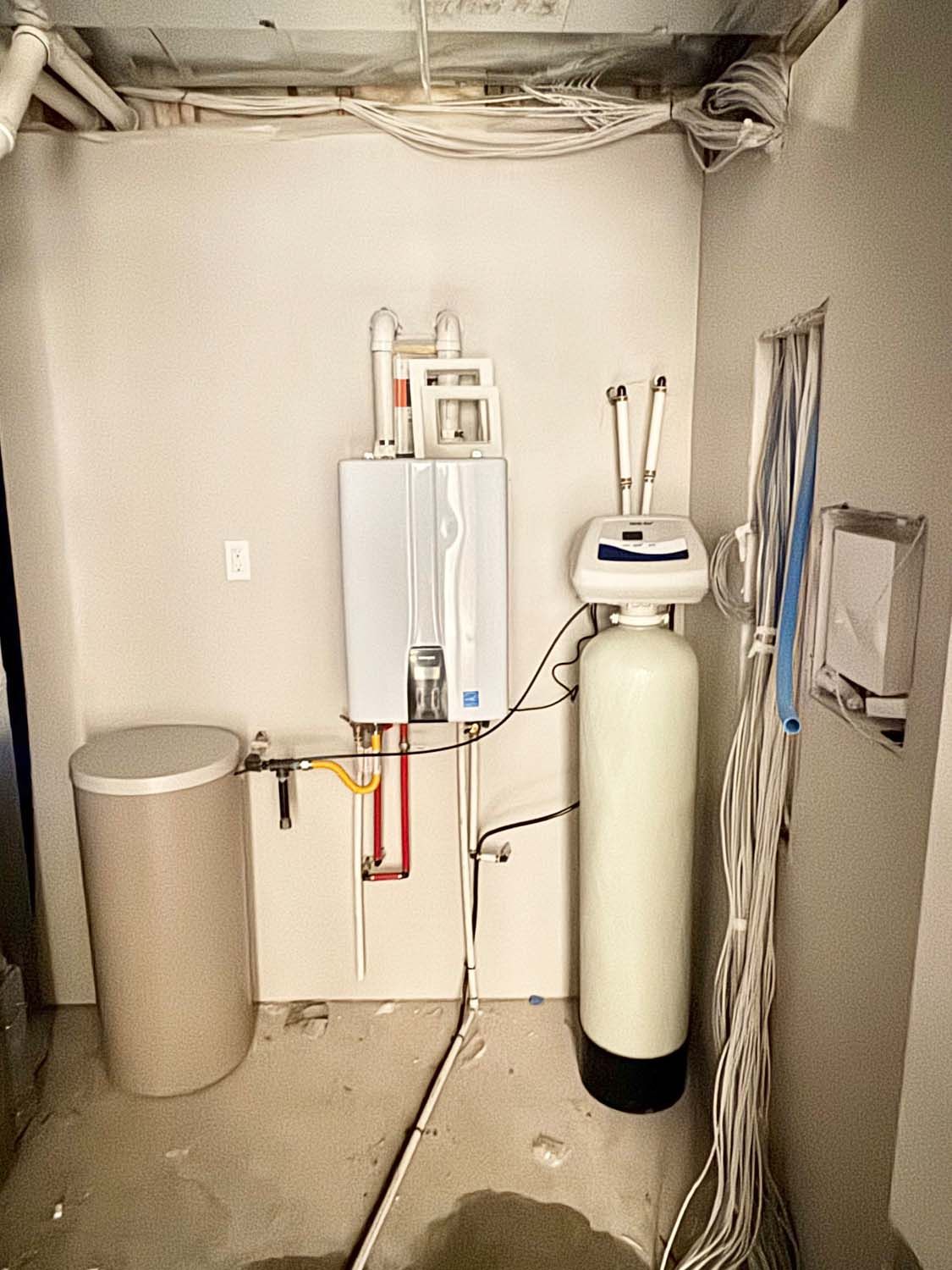 Water heater, water softener, and tank in unfinished utility room with exposed wires.