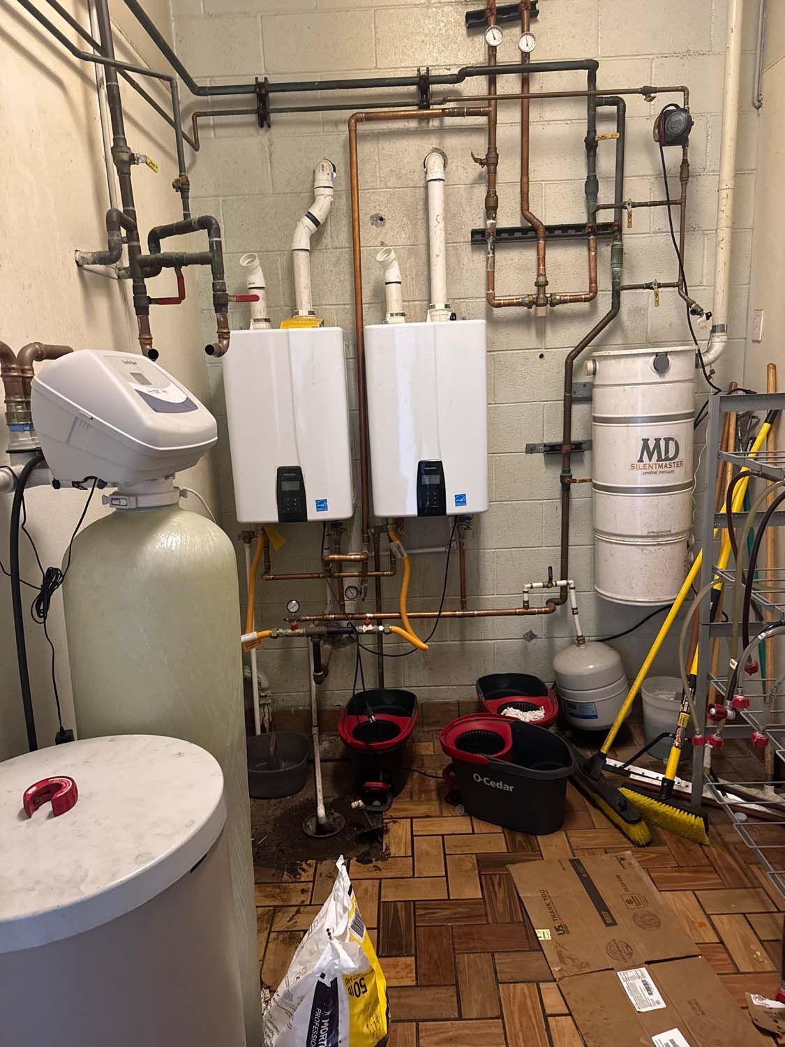 A utility room with water heaters, a water softener, and various pipes against a brick wall.