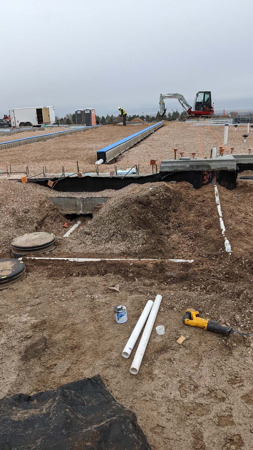 Construction site with exposed pipes and gravel. A worker operates heavy machinery in the background. Overcast day.