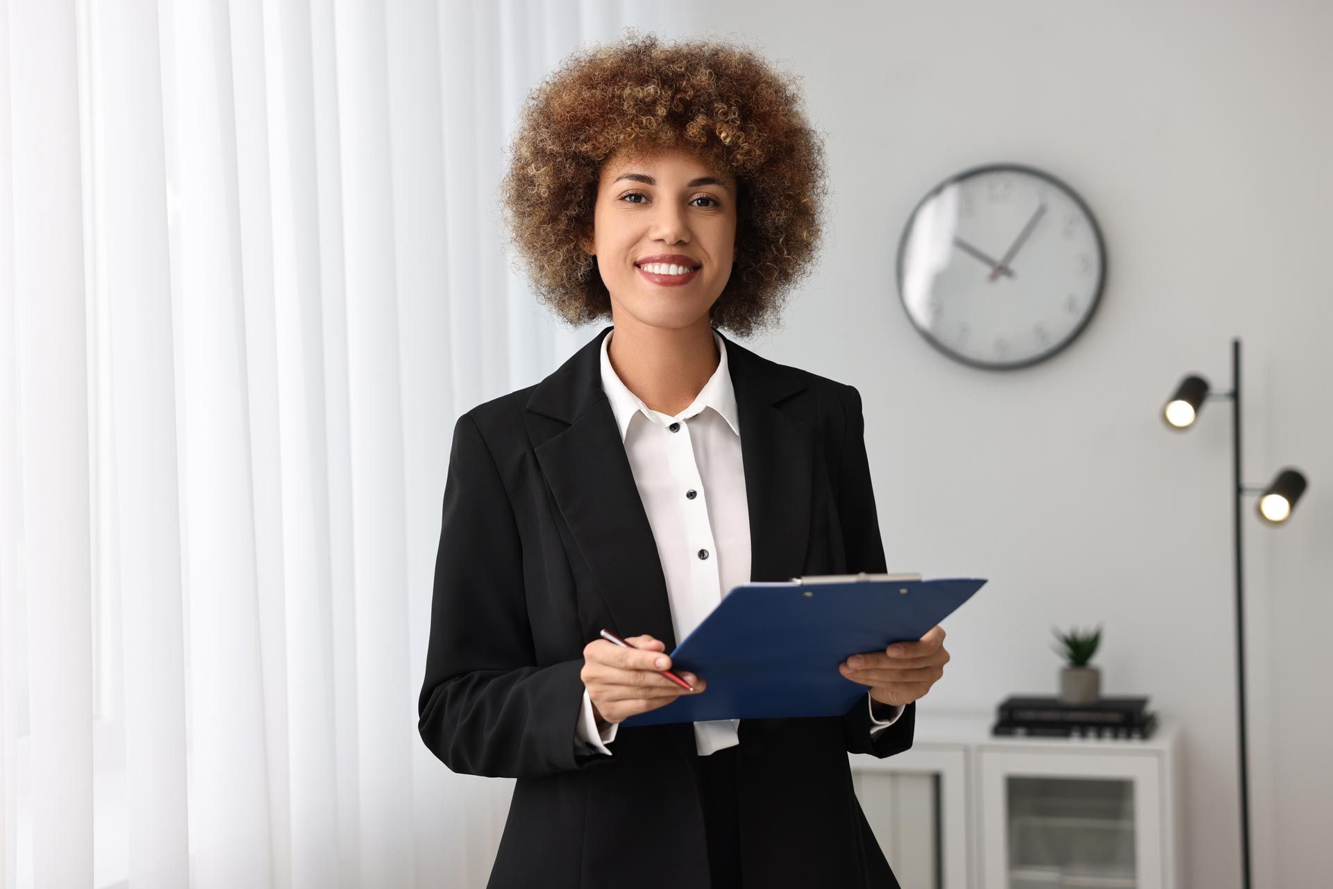 A woman in a suit is holding a clipboard and smiling