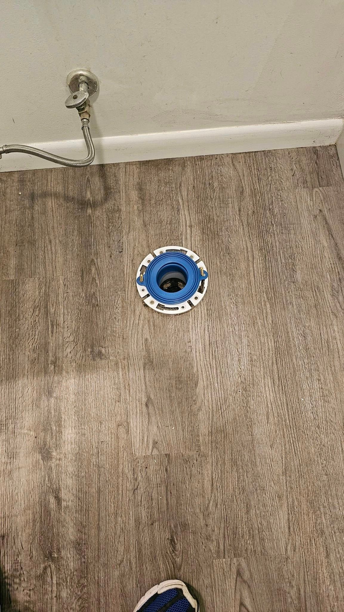 A close up of a toilet flange on a wooden floor in a bathroom.