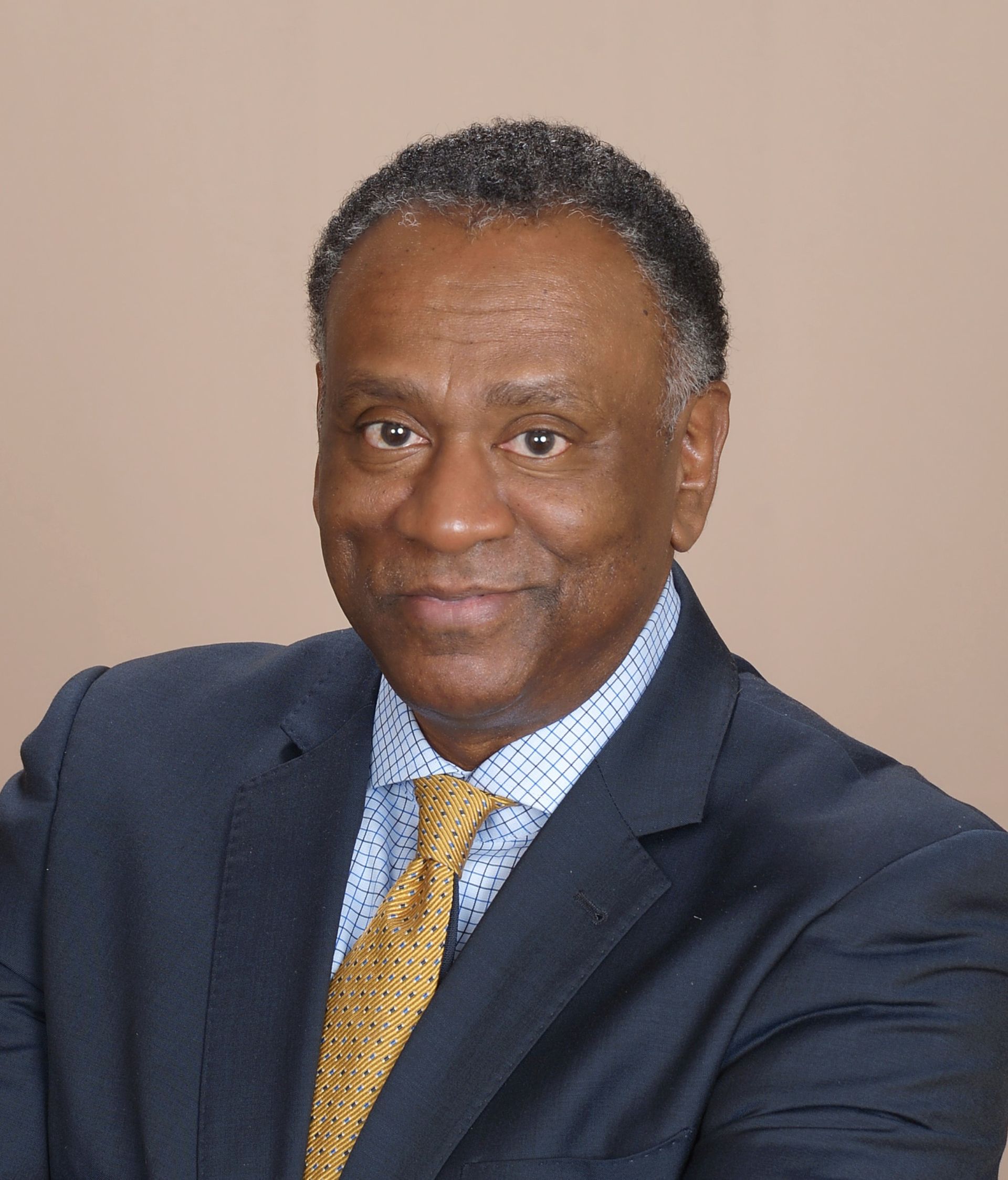 A professional portrait of a person in a blue suit, patterned light-blue shirt, and gold tie, smiling against a tan wall.