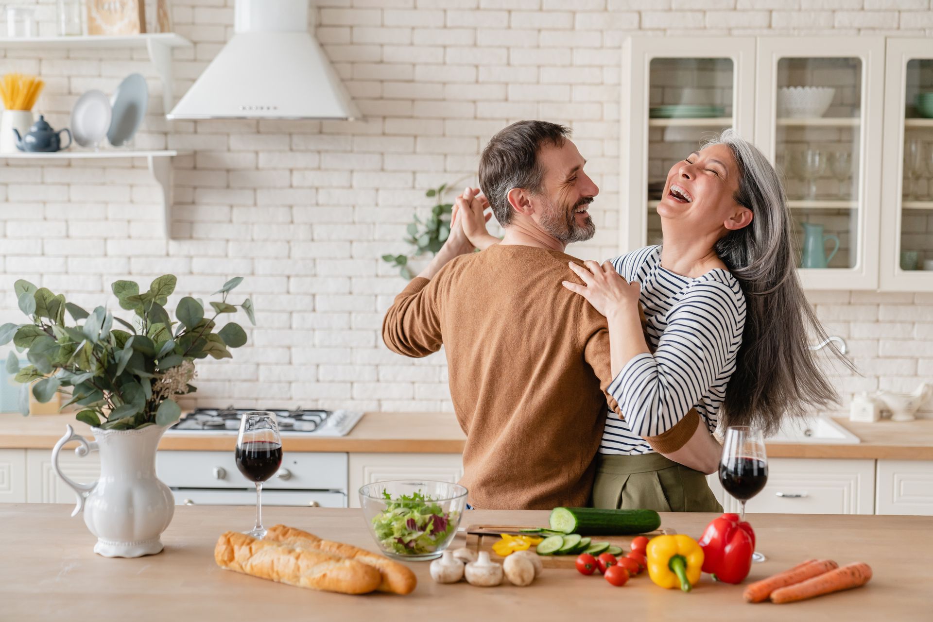 A couple dances joyfully in a bright, modern kitchen filled with fresh ingredients and glasses of red wine on the counter.