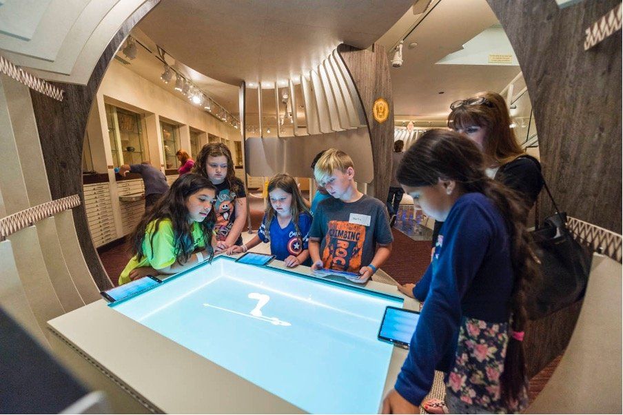 A group of children are standing around a table looking at a screen.