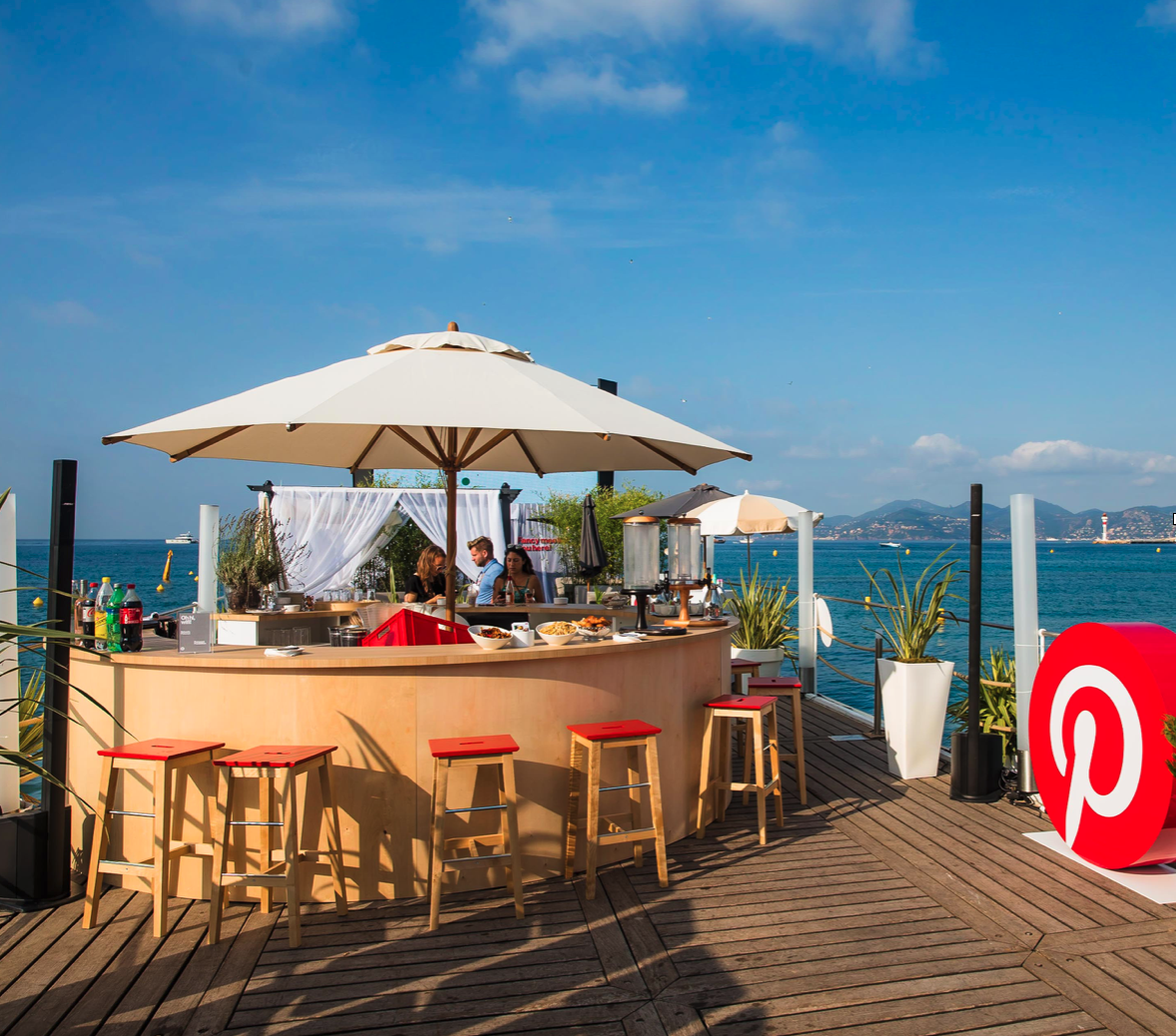 A bar with a large umbrella and stools next to the ocean