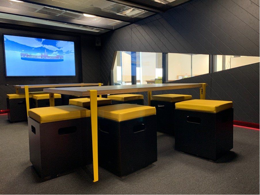 A room with tables and stools and a large screen on the wall