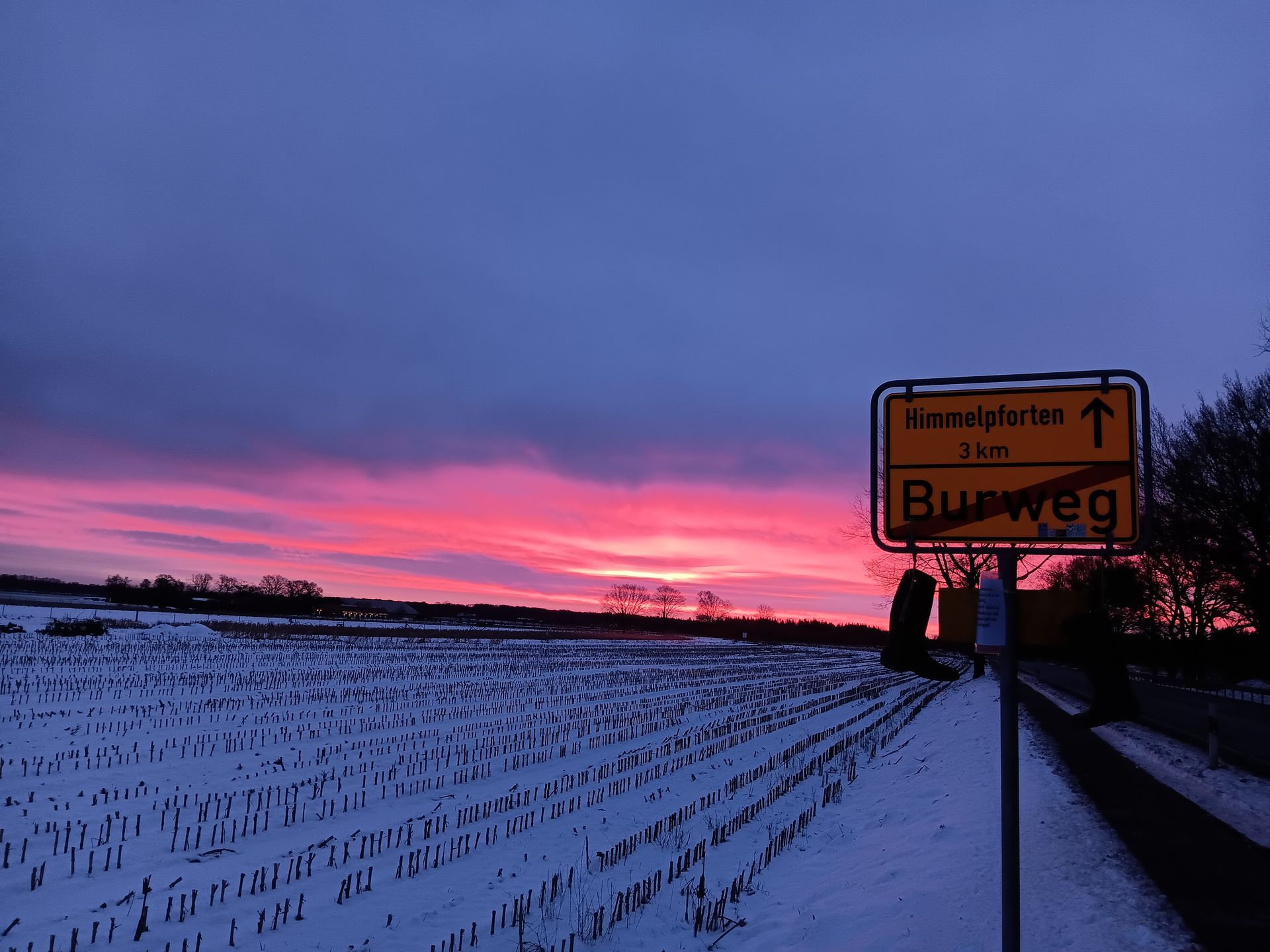 Sonnenuntergang über einem schneebedeckten Feld mit einem Schild „Burweg“. Der Himmel leuchtet rosa und violett.