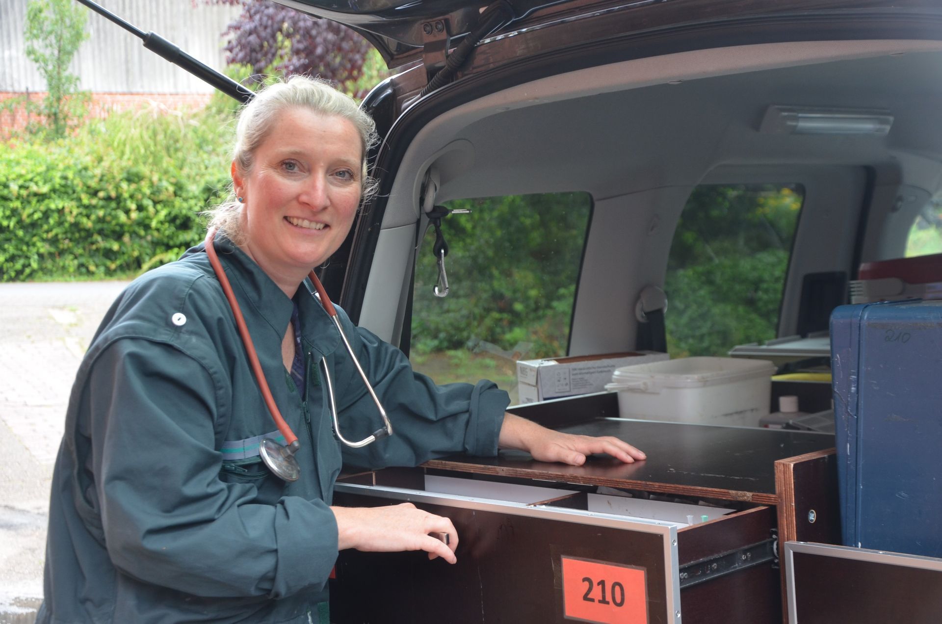 Tierärztin in grüner OP-Kleidung mit Stethoskop, auf der Ladefläche eines Fahrzeugs mit medizinischem Material.