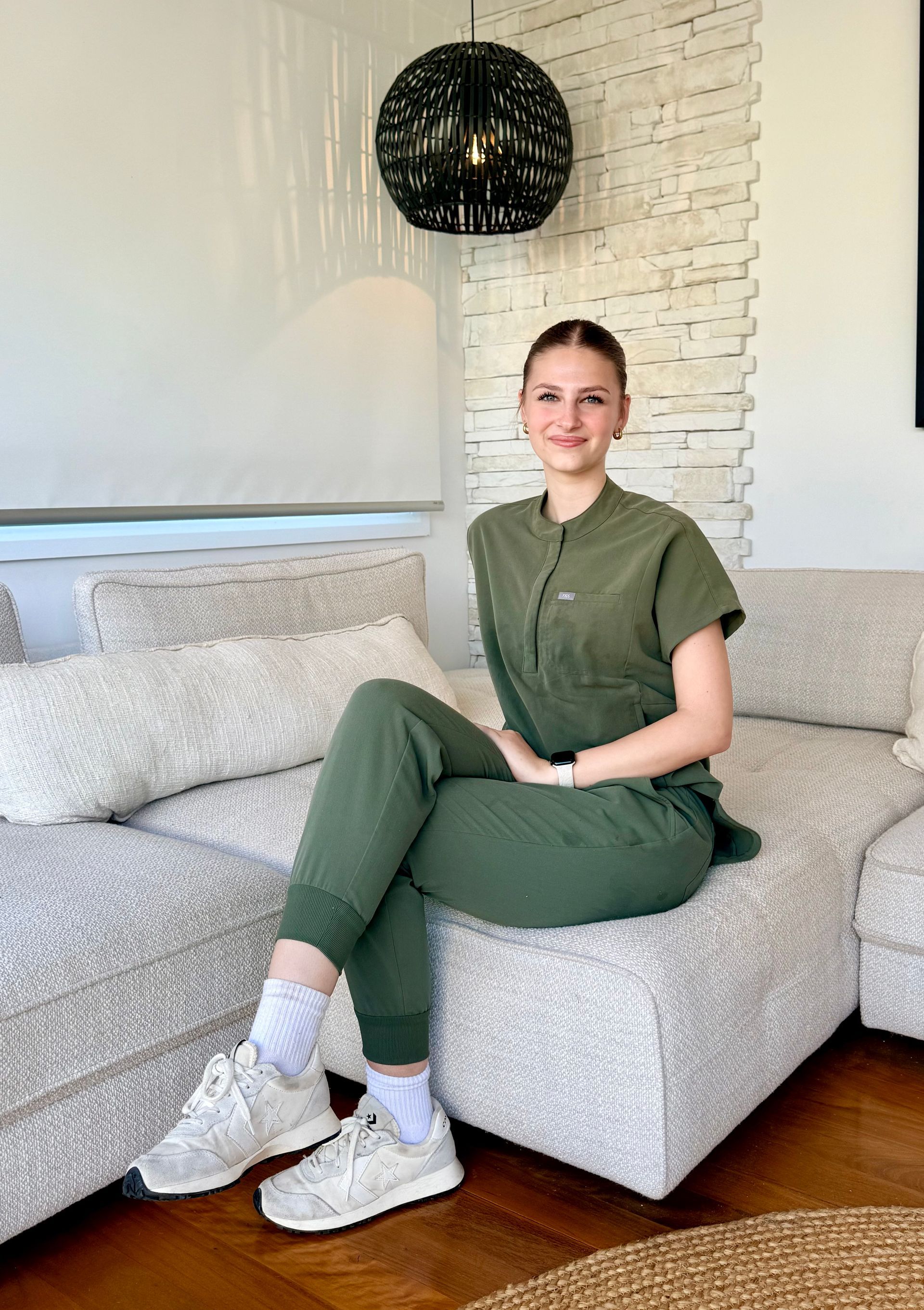 A smiling person wearing an olive green jumpsuit and white sneakers sits on a light-colored sectional sofa in a room.