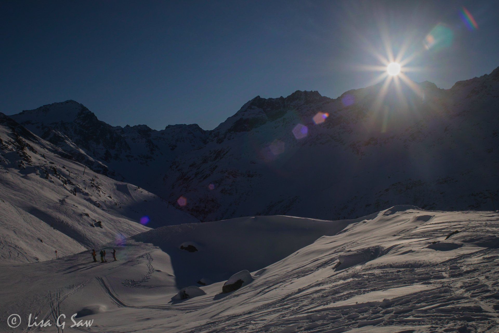 Sunburst behind snow capped mountains with skiers on piste