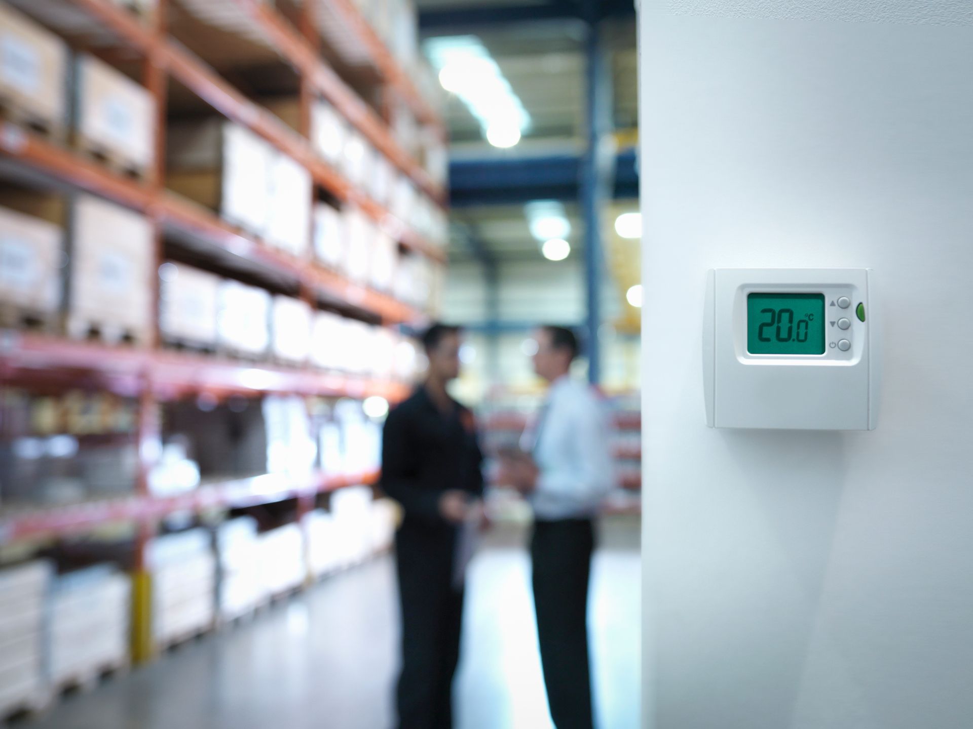 Two people are standing in a warehouse with a thermostat on the wall.