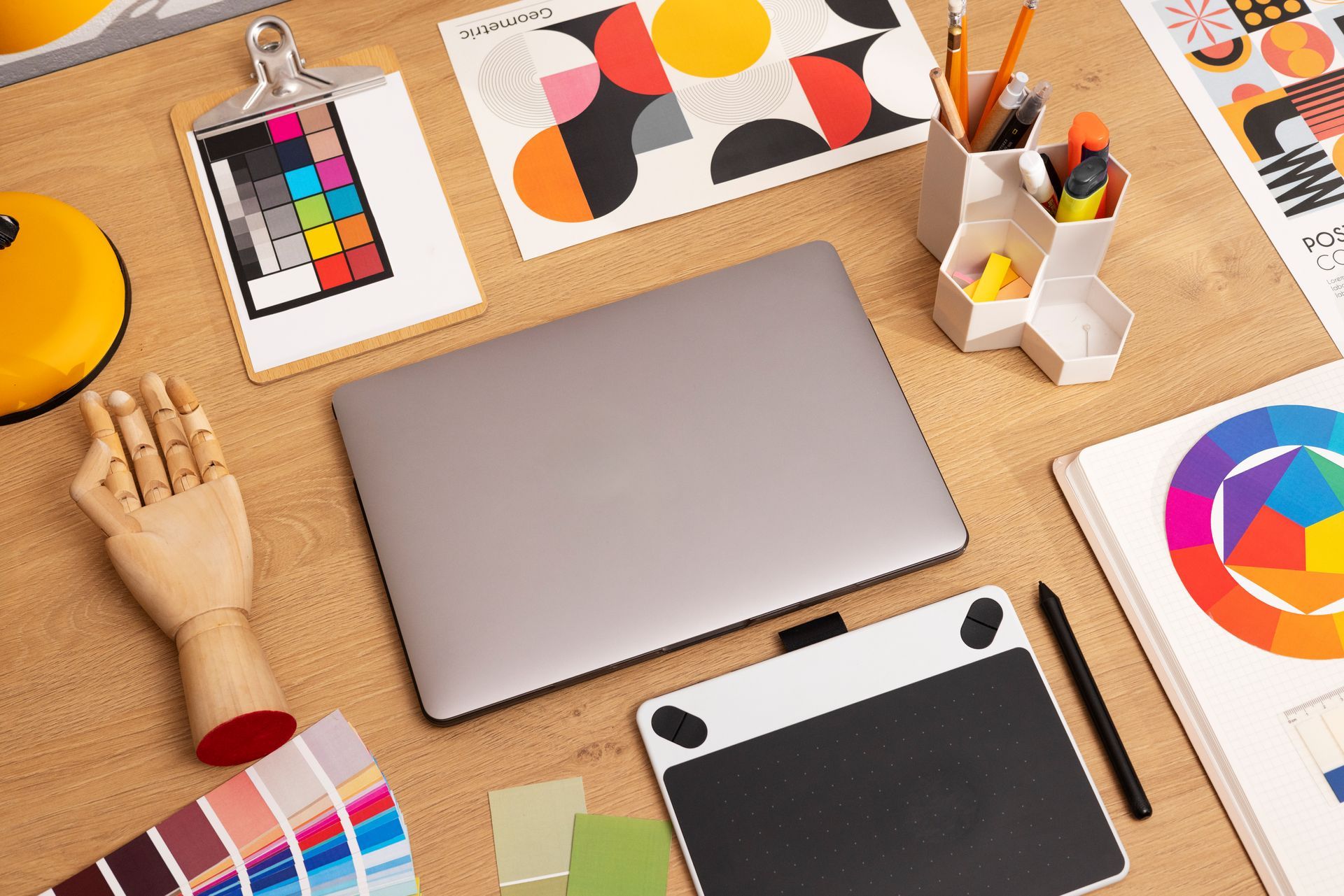 Computadora portátil, tableta gráfica y materiales de arte en un escritorio de madera, incluidas paletas de colores y maquetas de diseño.