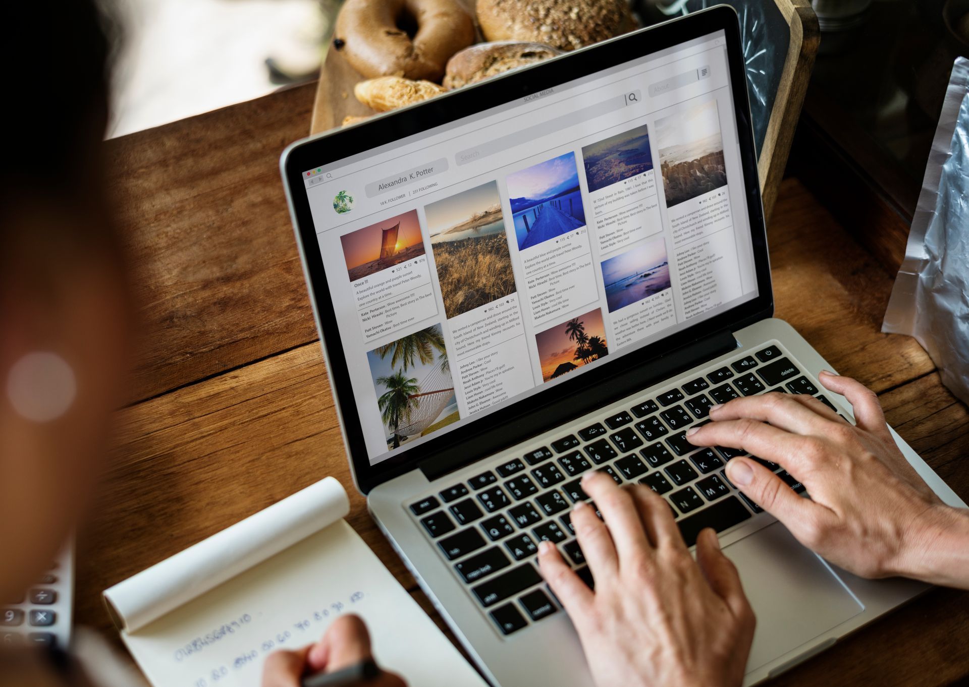 Persona escribiendo en una laptop, viendo imágenes en pantalla, con el cuaderno visible. Mesa de madera y bagels.