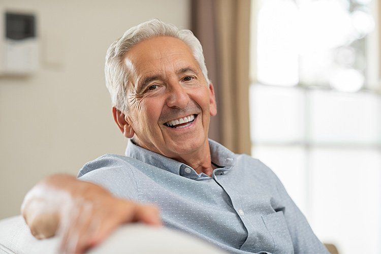 Smiling older man with white hair, light blue shirt, seated on a couch indoors.