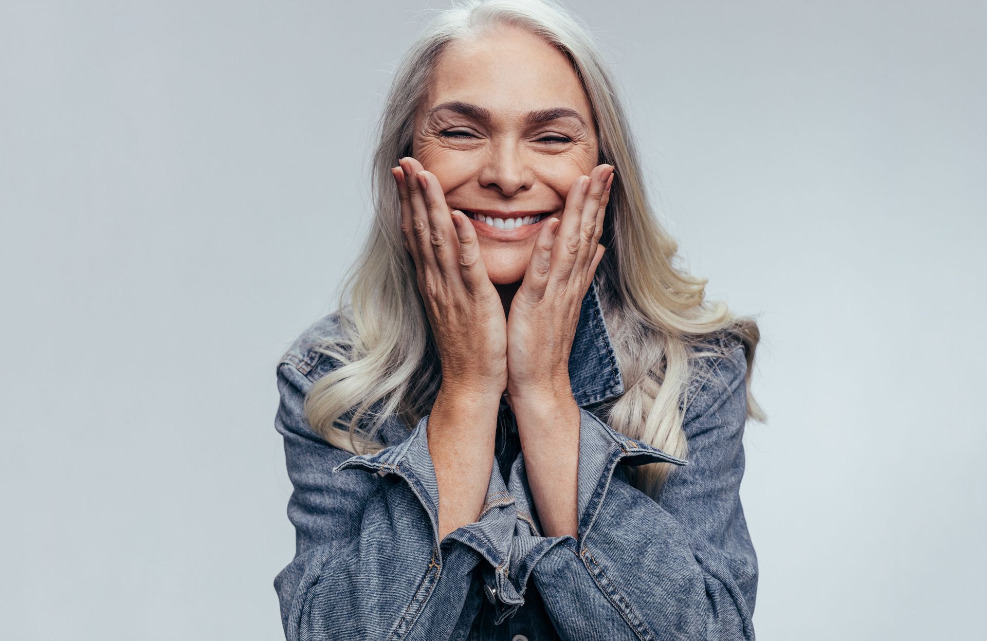 Smiling older woman with long gray hair, hands on her cheeks, wearing a denim jacket, against a neutral background.