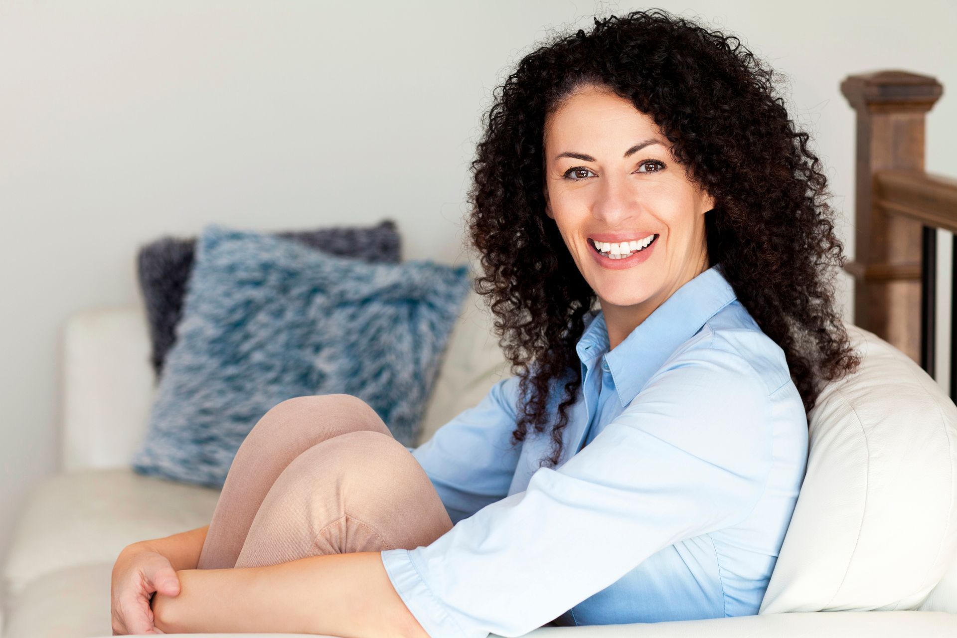 Woman with curly dark hair smiles, sitting on a white couch in a well-lit room, arms around legs.