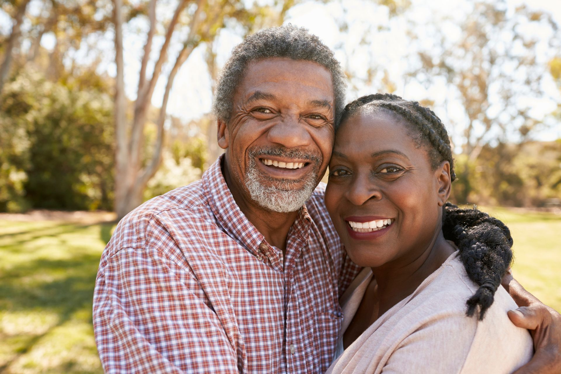 Smiling older Black couple embraces outdoors; man wears a checkered shirt, woman has braids.