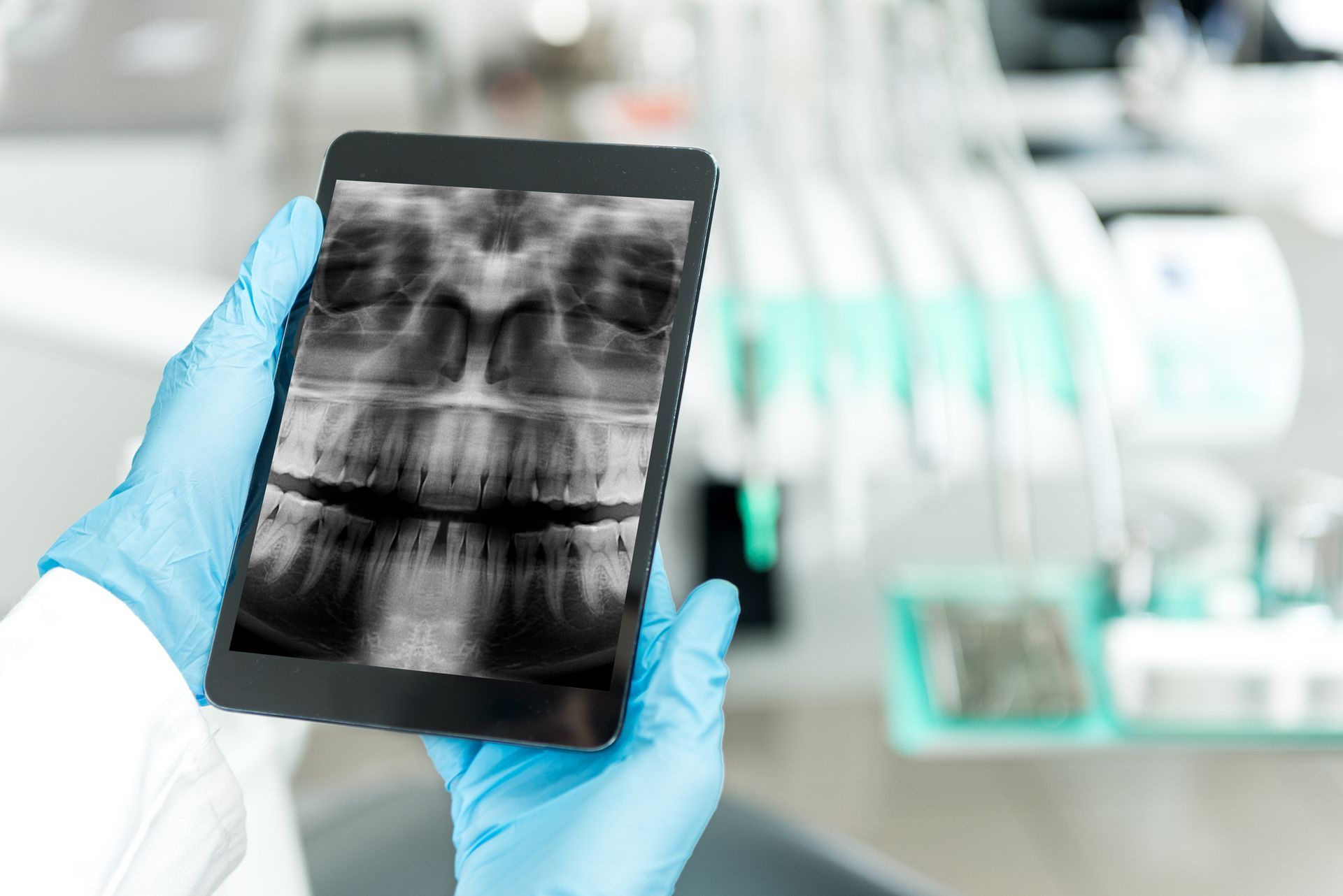 Dentist holding a tablet displaying a panoramic dental X-ray in a dental office setting. The dentist wears blue gloves and a lab coat.