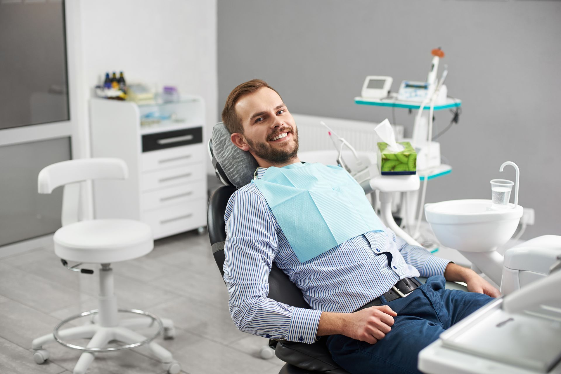 Man smiling in a dentist chair, wearing a blue paper bib. He is in a modern dental office setting.