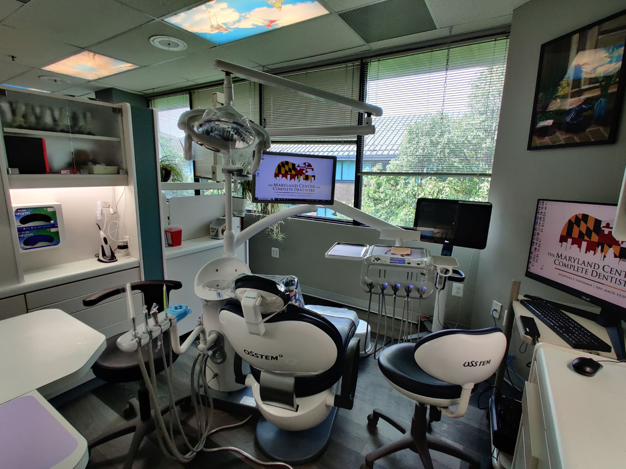 Dental exam room with chair, equipment, computer, and view of greenery through the window; the walls are a light blue with a cloudy sky accent on the ceiling.