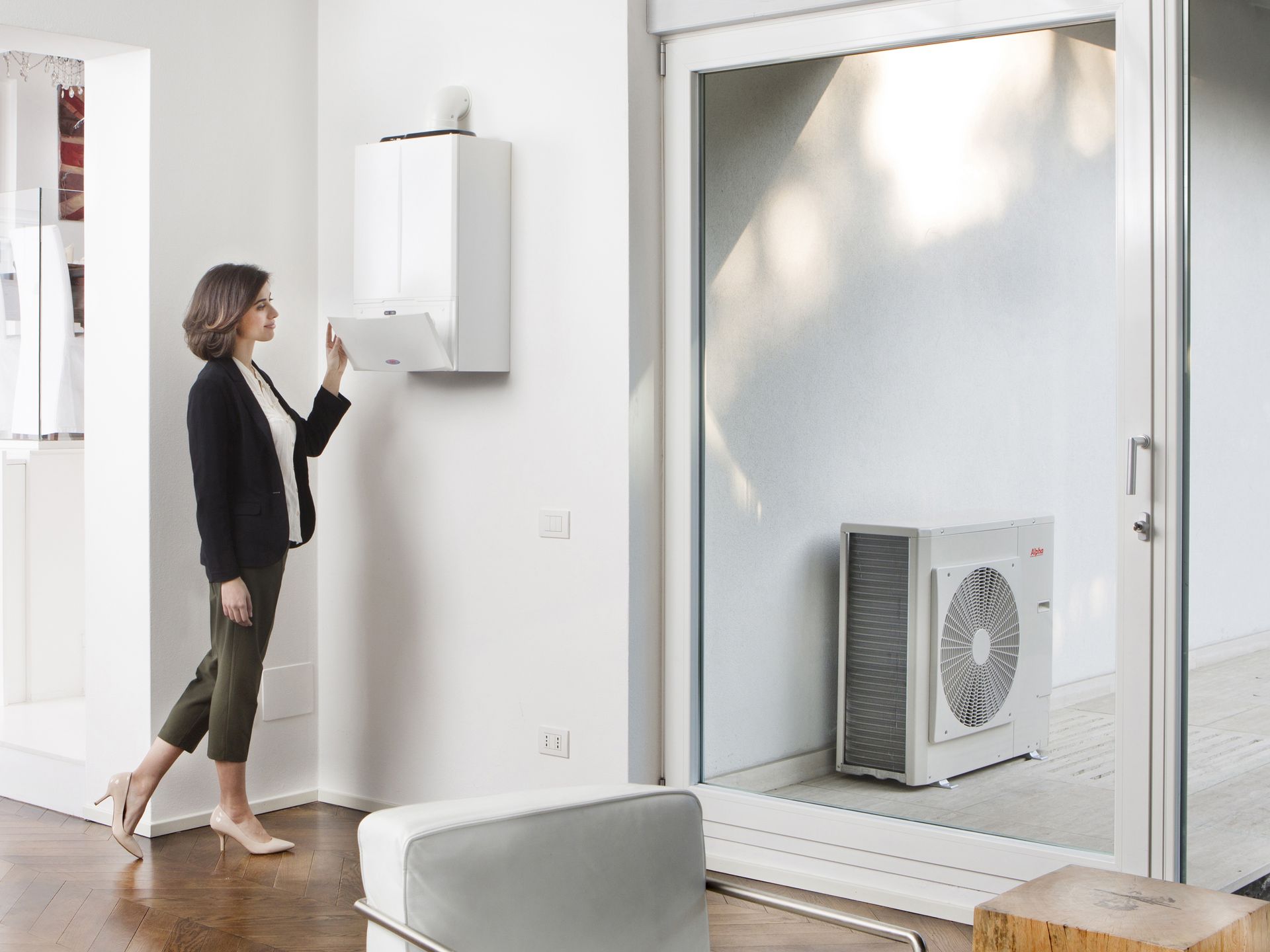 Woman interacting with wall-mounted heating unit in a modern white room; an outdoor unit is visible through a sliding door.