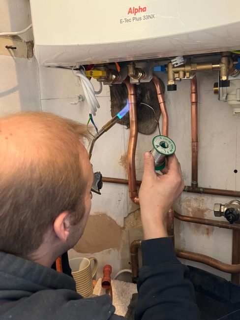 Plumber using a torch on copper pipes near a boiler in a room with exposed plaster.