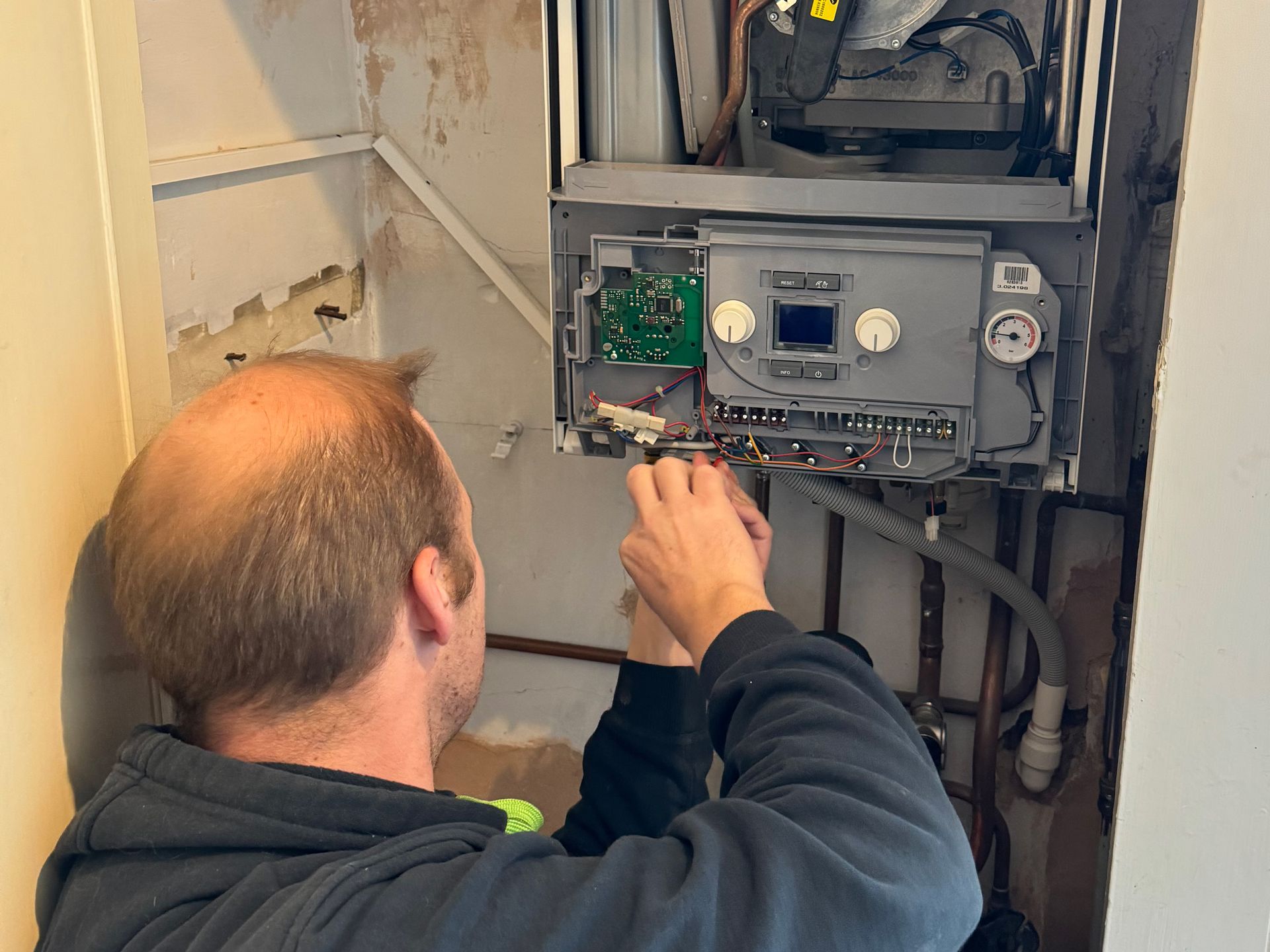 Man working on a boiler, inside an enclosure with exposed wiring, pipes.
