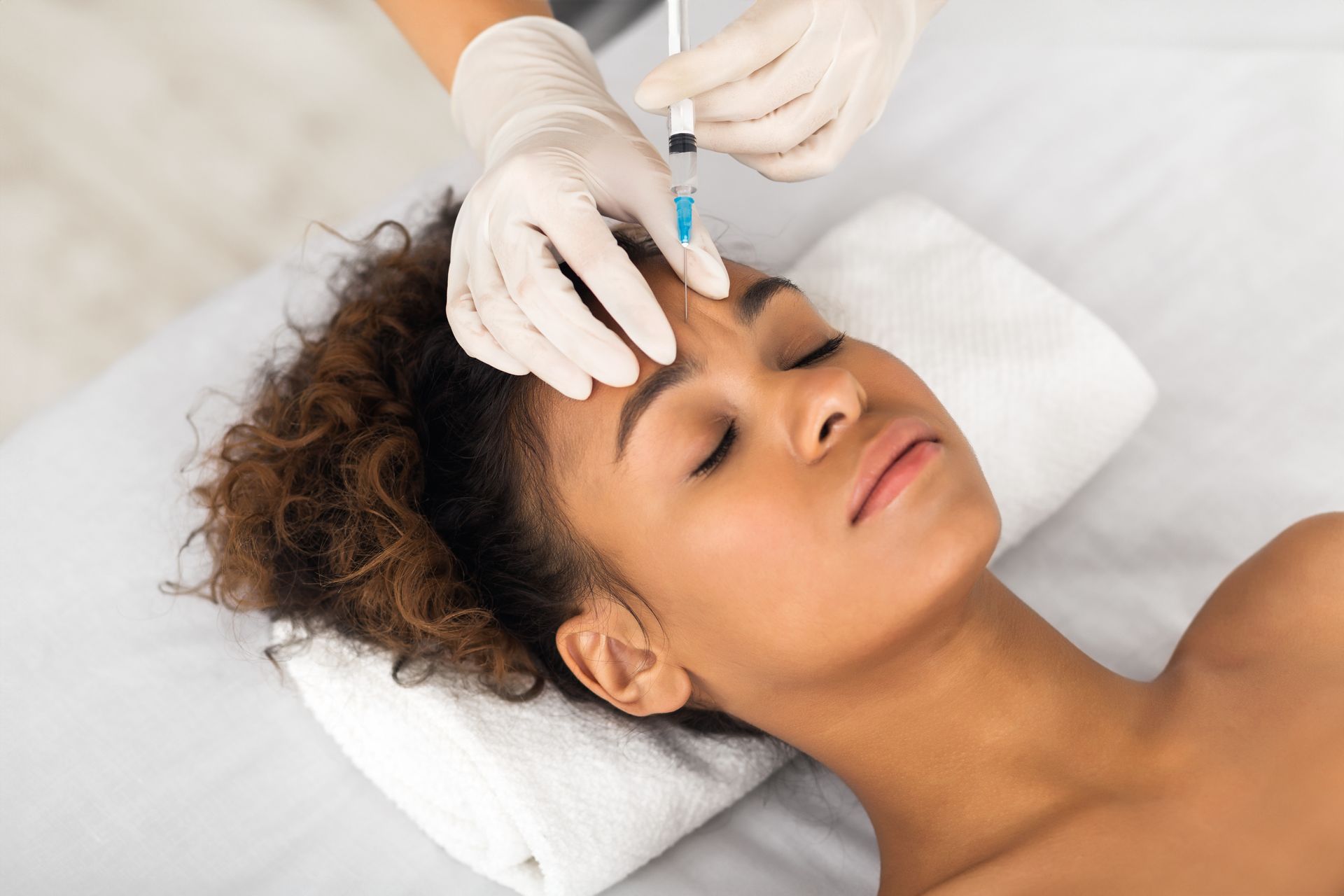 Woman receiving a Botox injection in her forehead, eyes closed, in a medical setting.