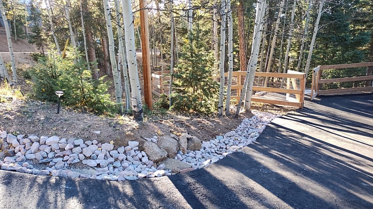 Asphalt driveway leads to a wooden gate in a forest setting, bordered by rocks and trees.