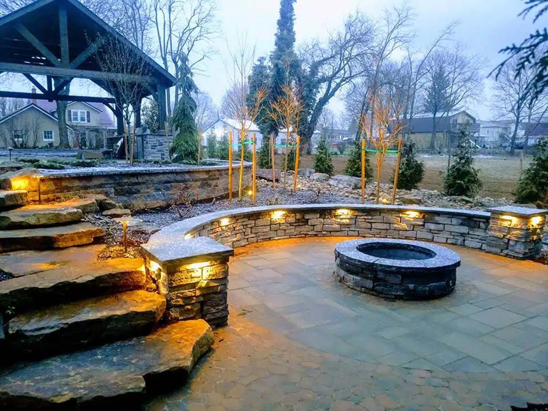 Stone patio with fire pit, steps, and retaining walls, illuminated by outdoor lights in a yard with a gazebo.