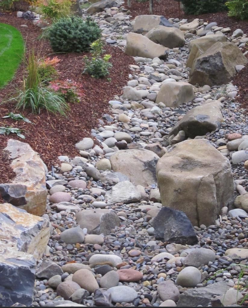 Dry creek bed landscaping with large rocks, pebbles, and mulch. Green and orange plants border the creek.
