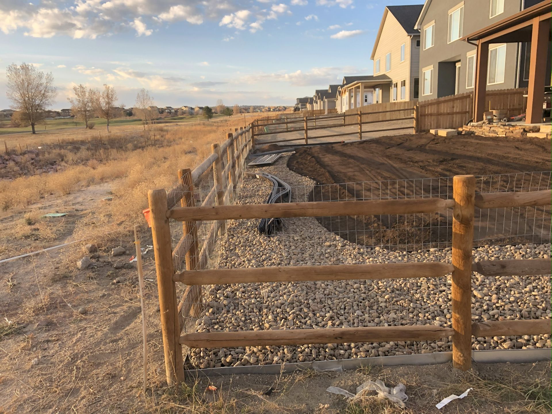 A wooden fence surrounds a gravel area with houses in the background