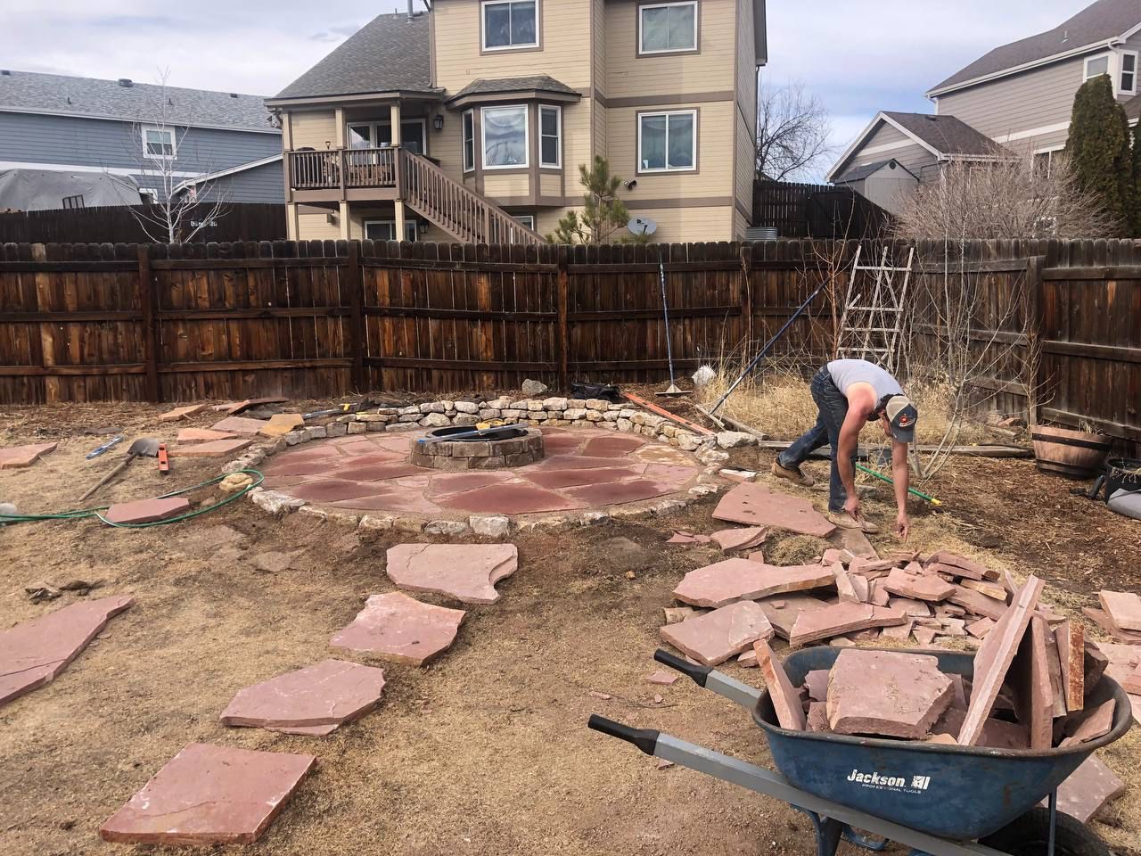 A man is working in a backyard with a wheelbarrow full of rocks.